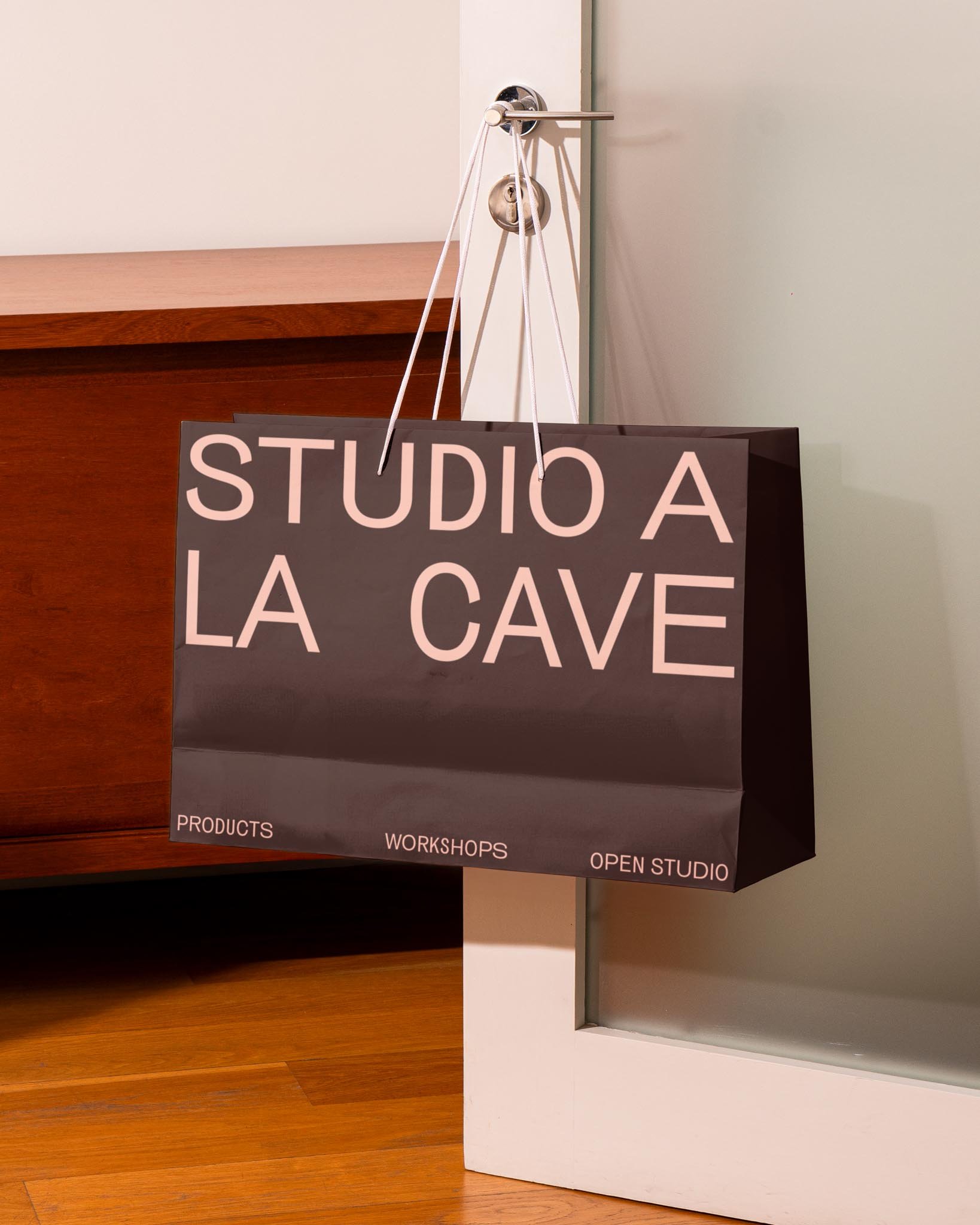 Black paper shopping bag with pink text that reads, 'STUDIO A LA CAVE,' hanging from a door handle, with 'PRODUCTS,' 'WORKSHOPS,' and 'OPEN STUDIO' printed at the bottom.