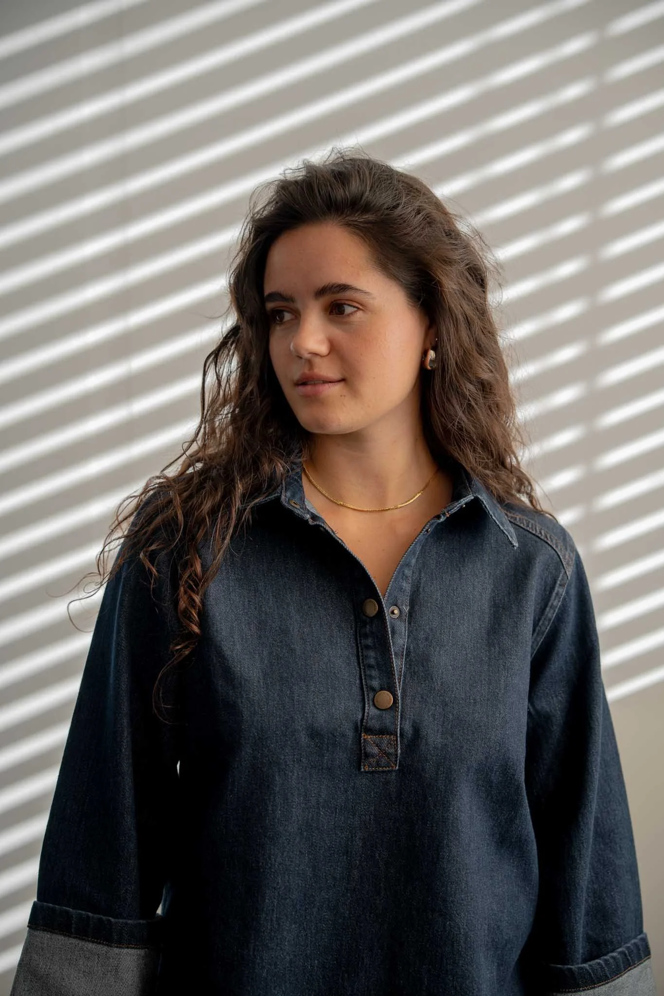 A young woman with long, wavy brown hair wearing a dark denim shirt and gold jewelry, standing in front of a window with striped blinds.
