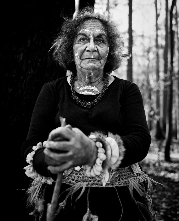 An elderly woman with traditional face paint, wearing jewelry and bracelets, holding a walking stick, standing in a forest.