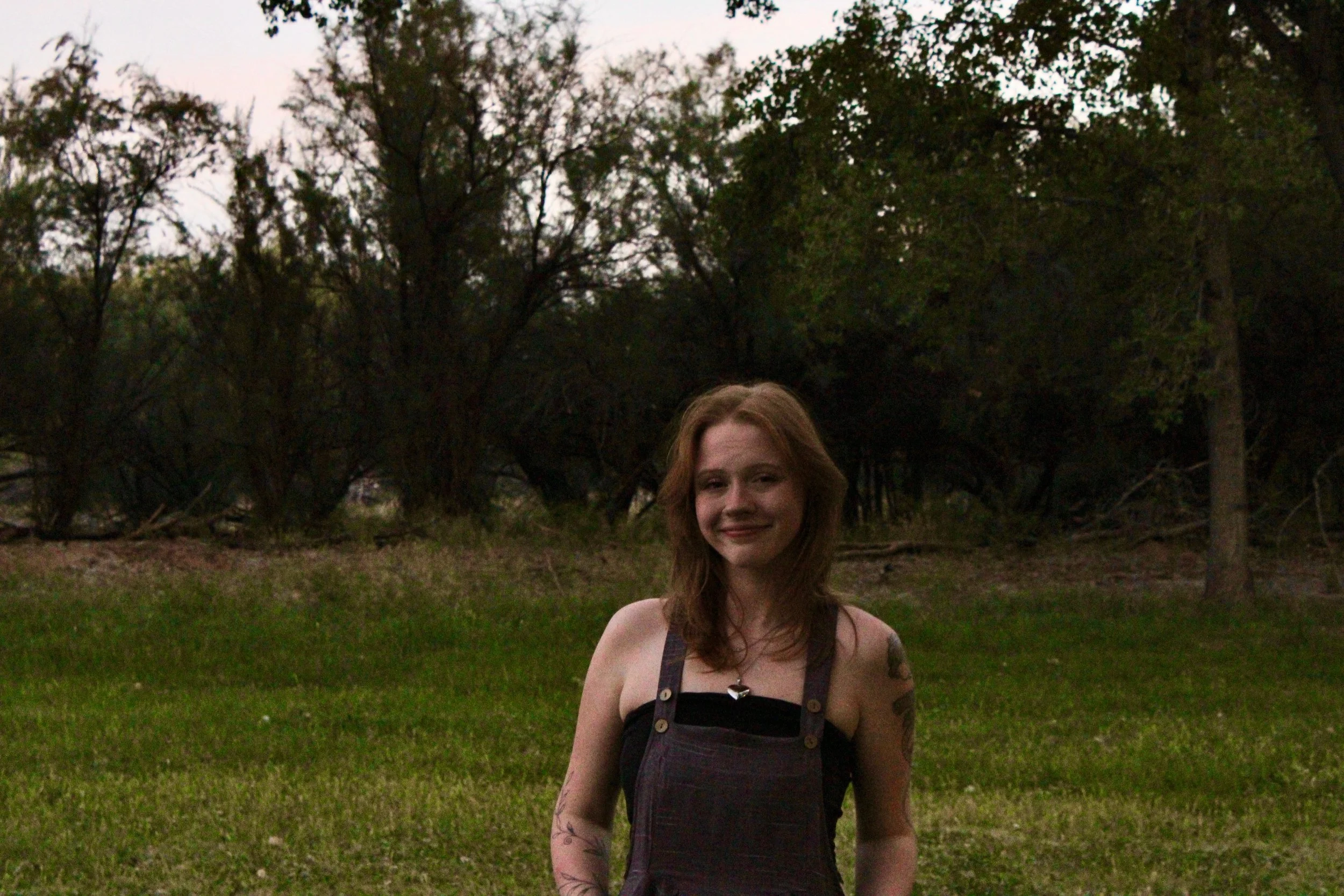 A young woman with long reddish hair standing outdoors in a grassy area, wearing a black dress with suspenders, in front of trees with a cloudy sky overhead.