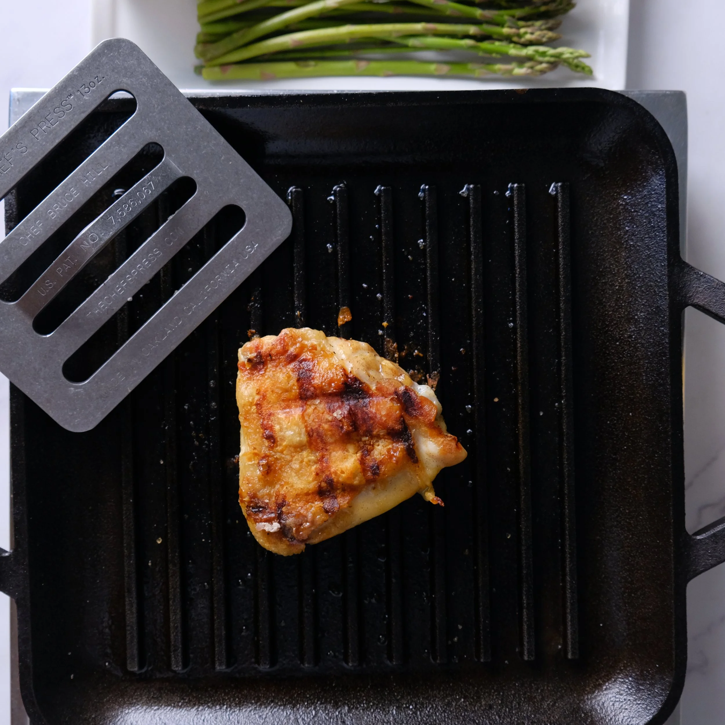 A cooked chicken thigh on a grill pan with a grill press nearby, with green asparagus stalks in the background.