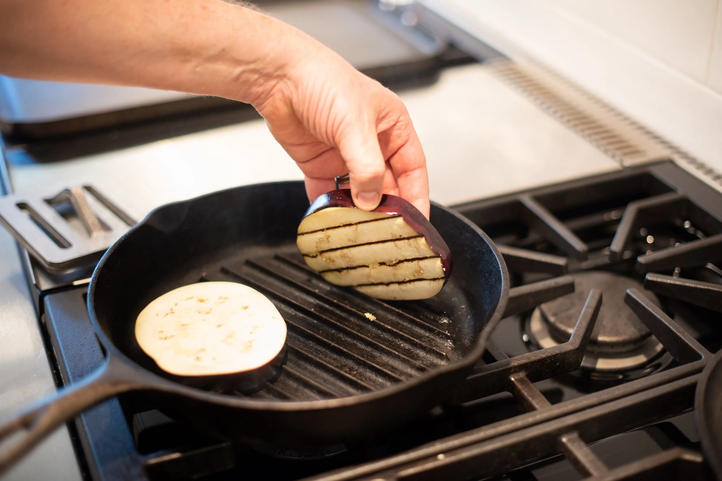 Person cooking sliced eggplant and sliced eggplant in a frying pan on a stove.
