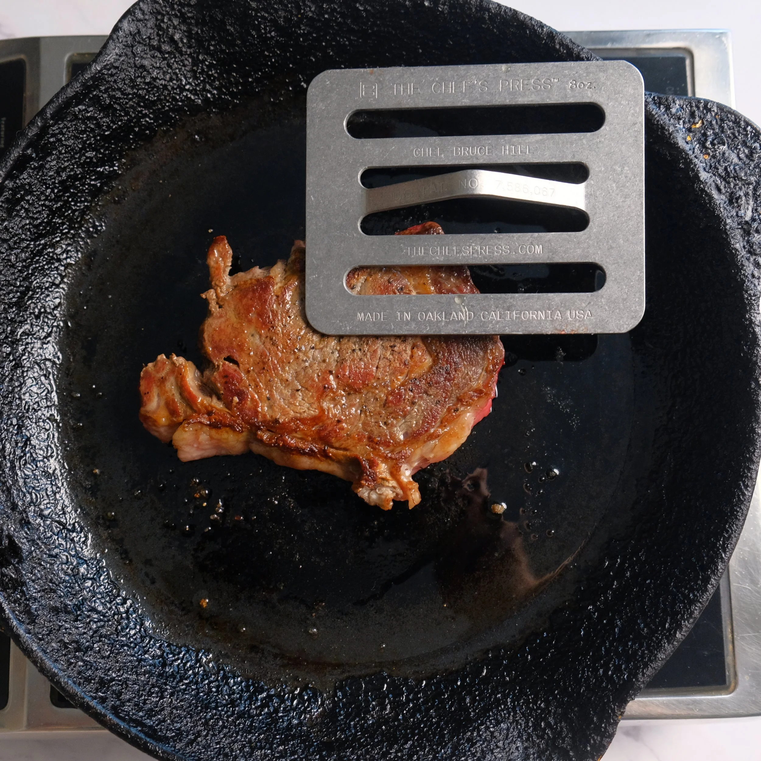 A cooked steak on a cast iron skillet with a metal grill resting on top that reads 'THE CHEF'S PRESS', 'GILL BRUCE HILL', 'MADE IN OAKLAND CALIFORNIA USA', and the website 'THECHEFSPRESS.COM'.