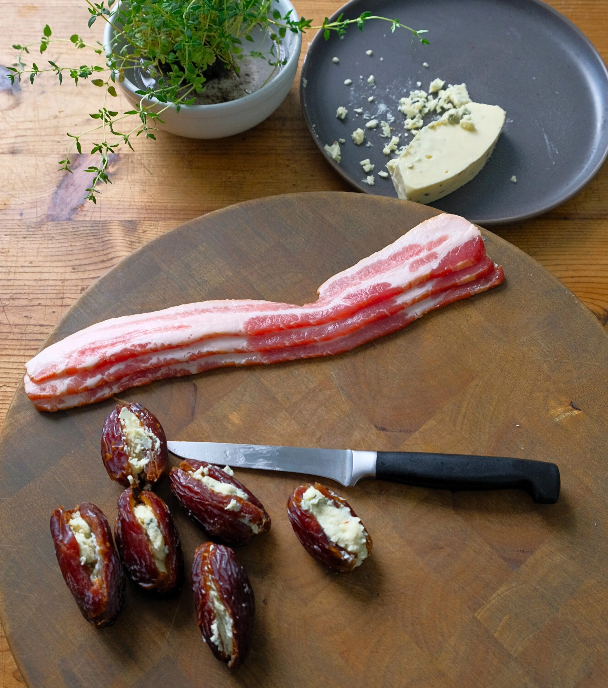 A wooden table with a cutting board holding slices of raw bacon, stuffed dates with cheese, a knife, a plate of blue cheese, and a potted herb plant.