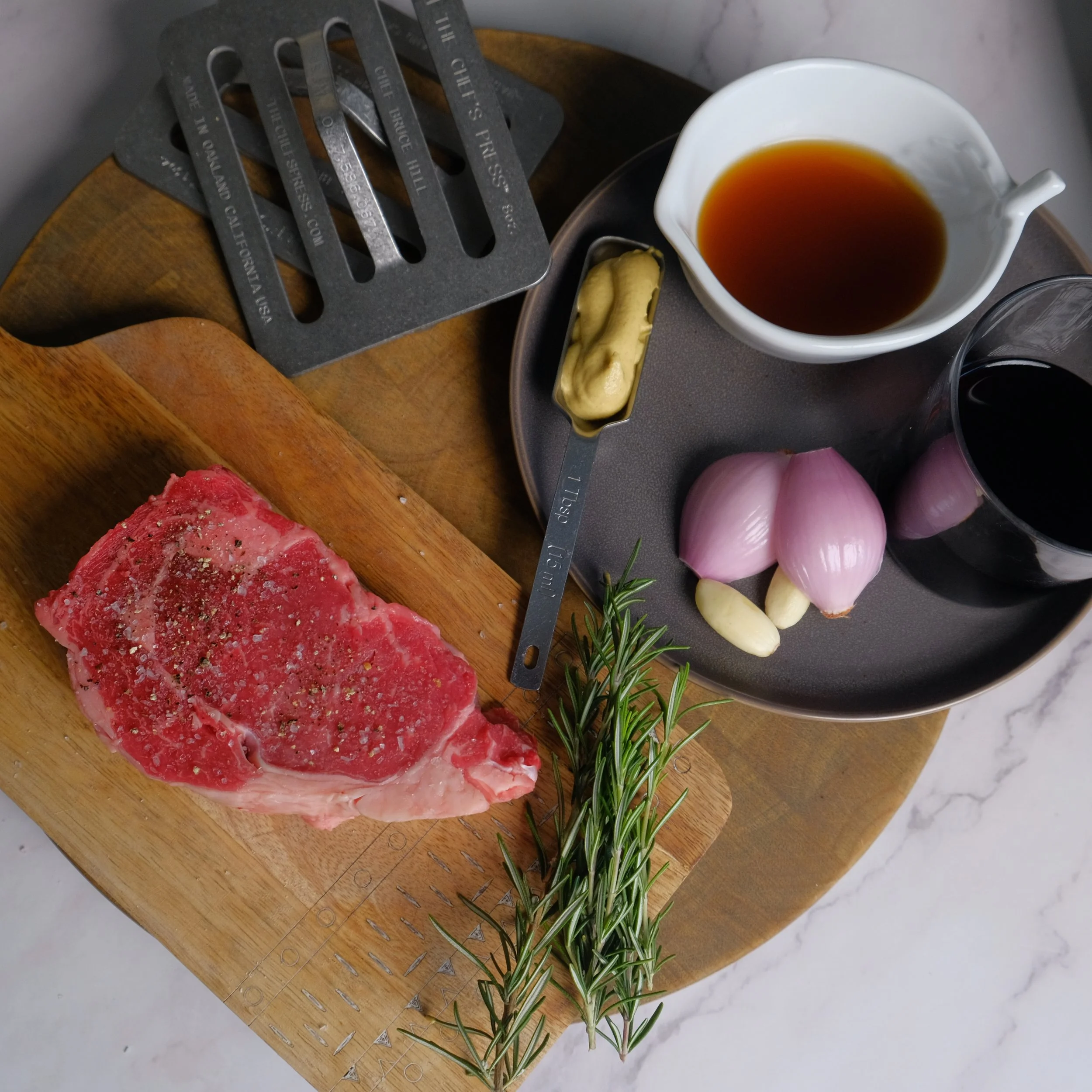 Raw ribeye steak seasoned with salt and pepper, with sprigs of rosemary on a wooden cutting board. Nearby are two whole garlic cloves, three shallots, and a small scoop of mustard. On a tray, there are bowls of black coffee, soy sauce, and a white bo