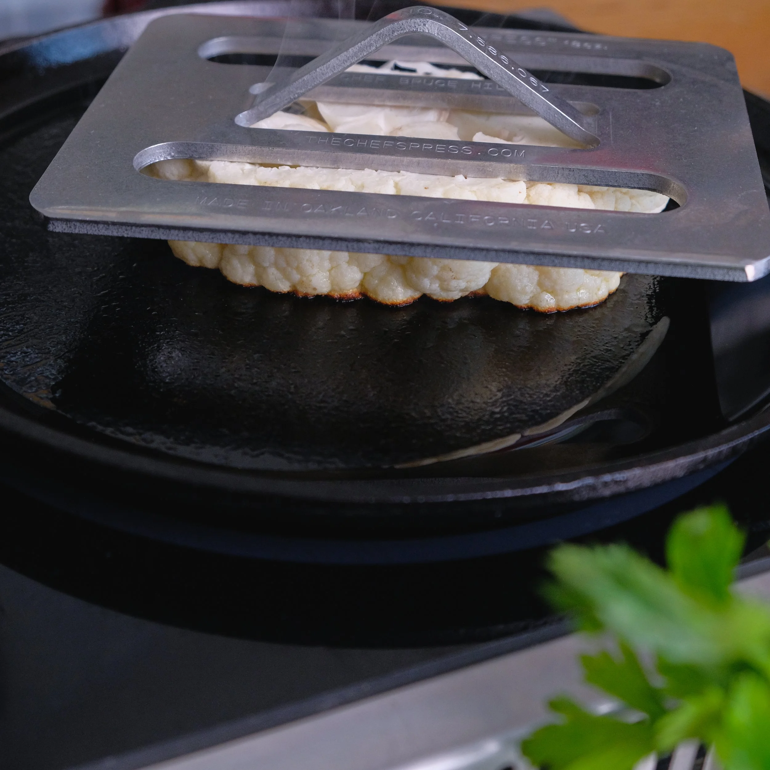 A cauliflower head is being pressed on a heated griddle with a metal press in a kitchen setting.