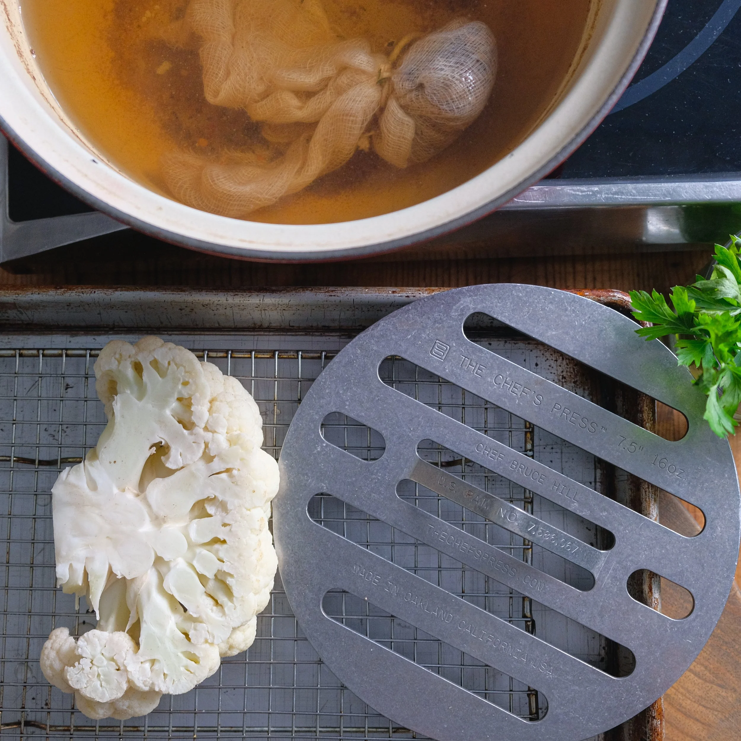 Raw cauliflower and a metal grill tray, with a pot of broth with a tea bag and herbs on a stovetop.