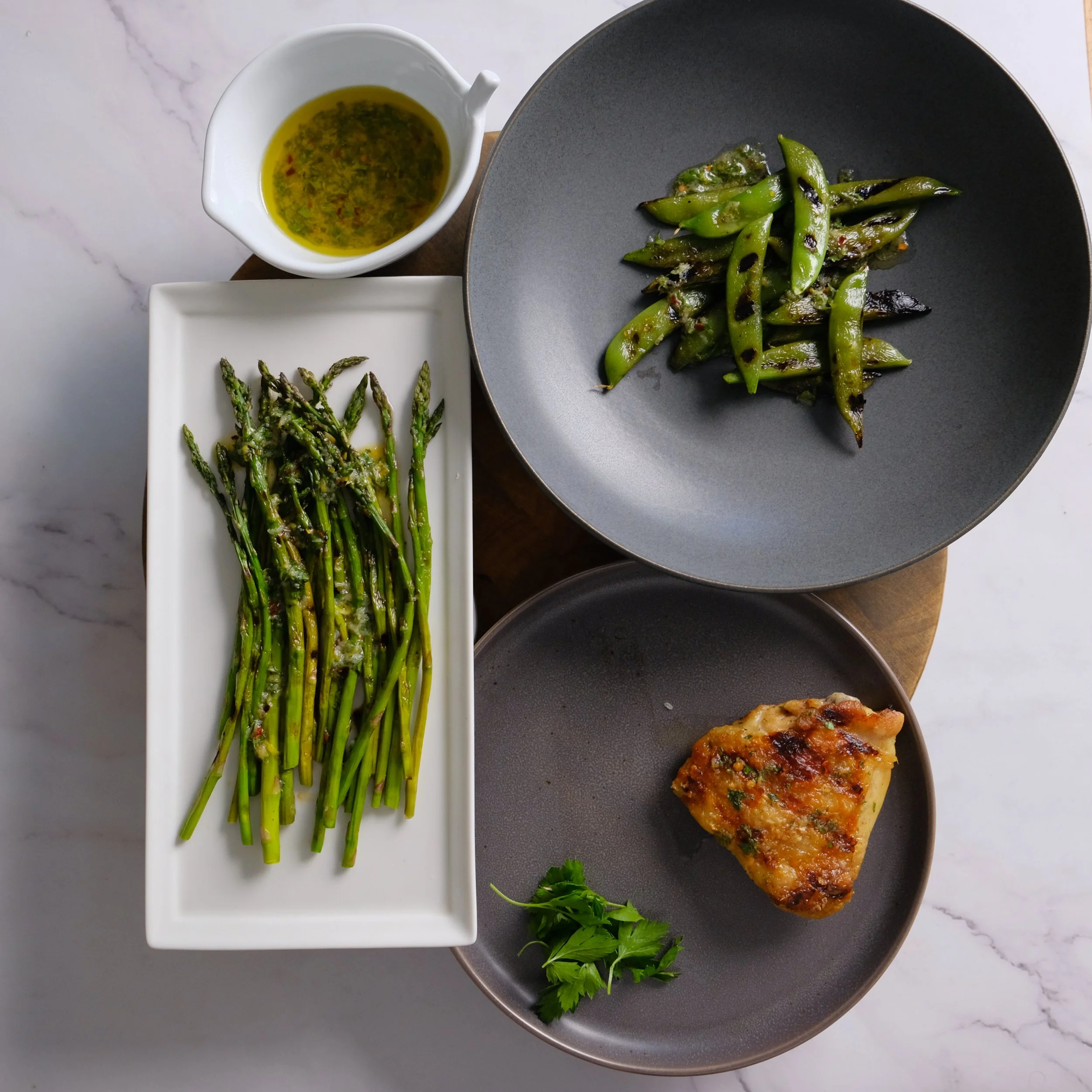 A plate of grilled green beans with charred marks, a bowl of grilled asparagus with garlic, a portion of baked lasagna with cheese and herbs, and a small container of olive oil with herbs, all arranged on a white marble surface.