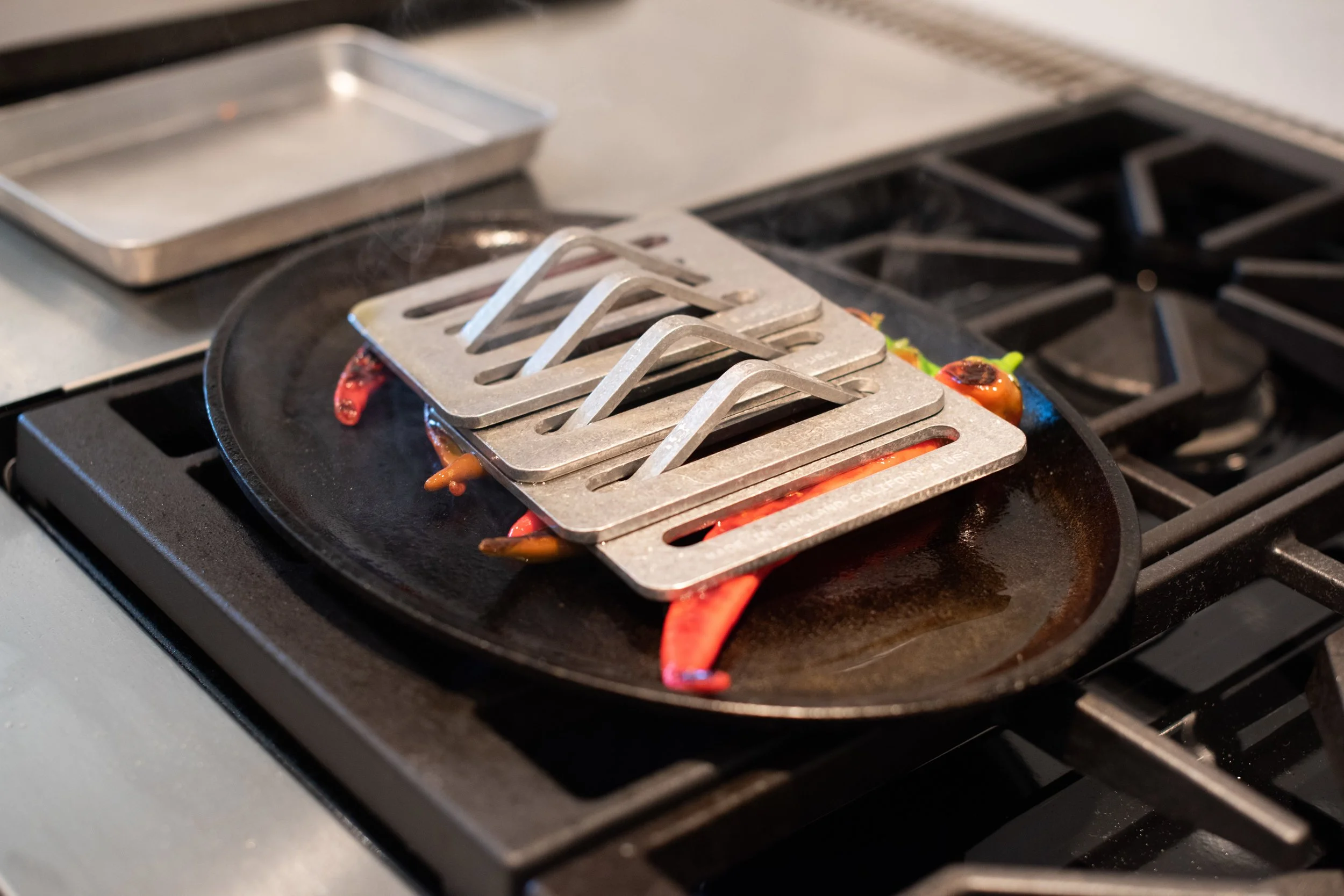 A stovetop with a cast iron skillet covering colorful chili peppers, and metal grill toppers placed on the peppers.