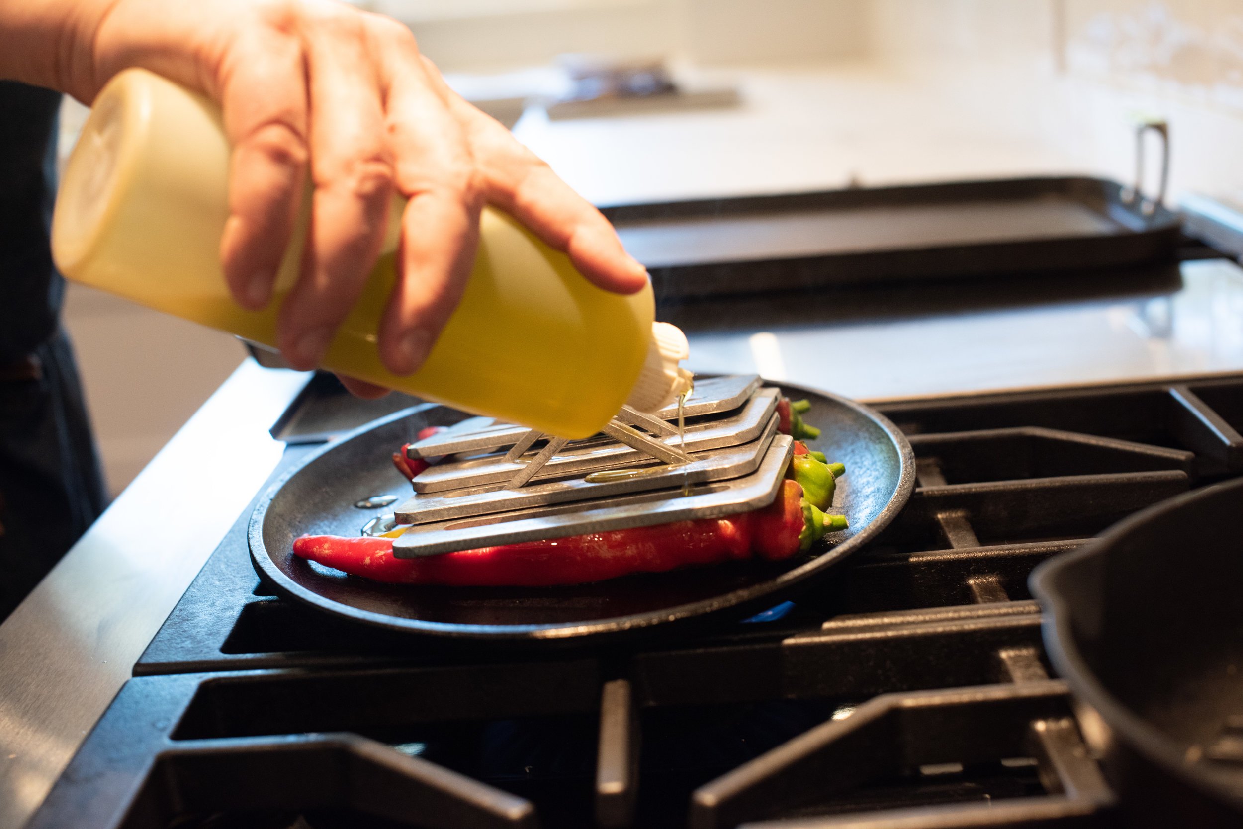 Someone pouring yellow cooking oil onto a skillet with peppers and metal molds on a stove.