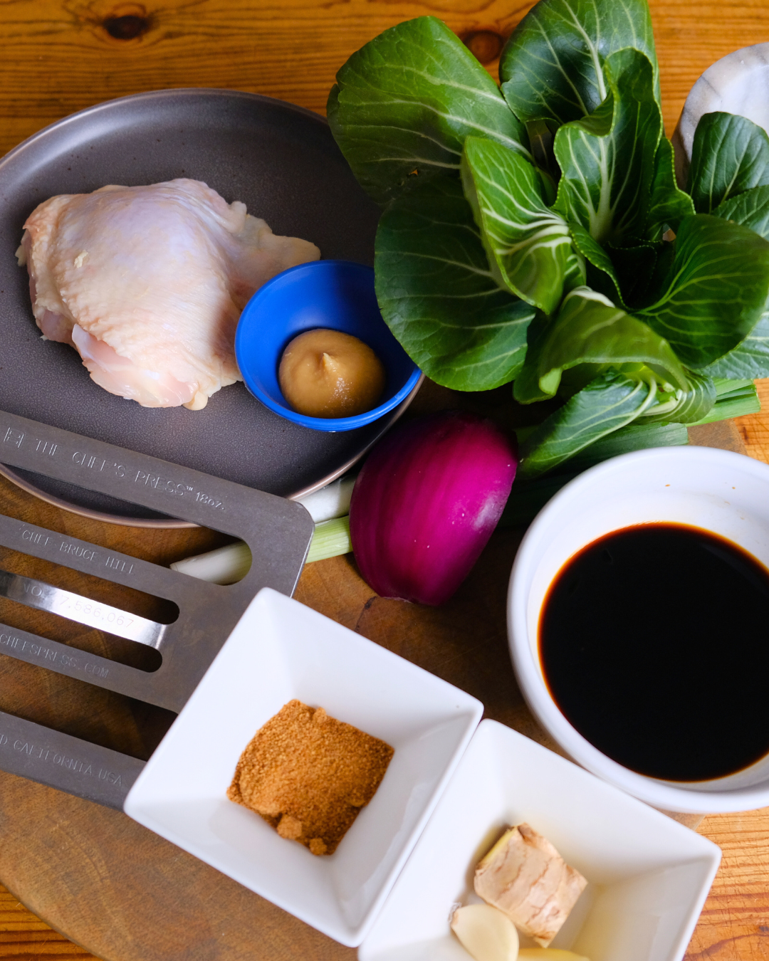 Ingredients for cooking, including chicken thigh, mustard, leafy greens, red onion, soy sauce, garlic, ginger, and seasonings.