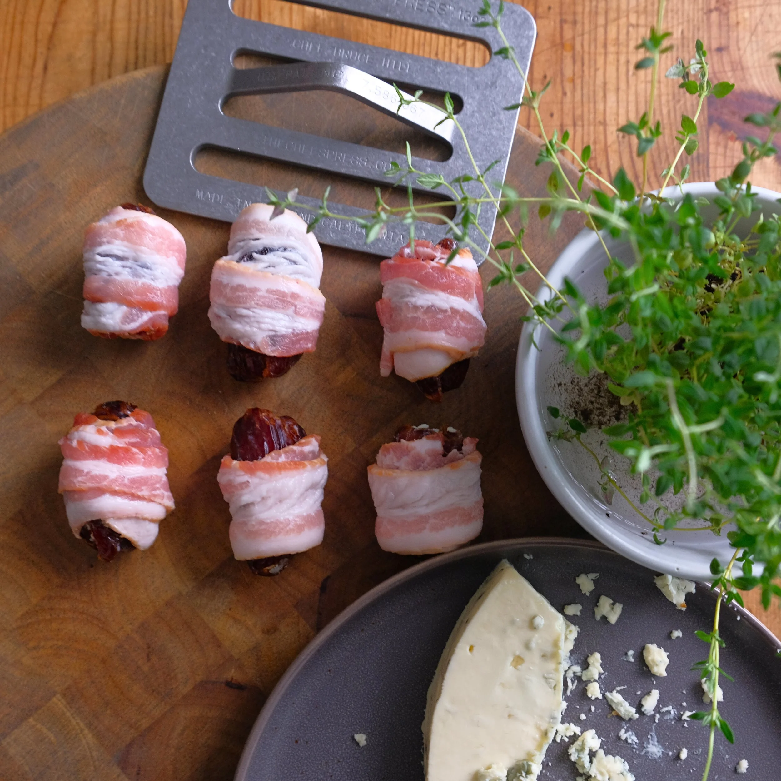 Bacon-wrapped dates on a wooden cutting board, a plant in a white pot, and a wheel of cheese on a gray plate.