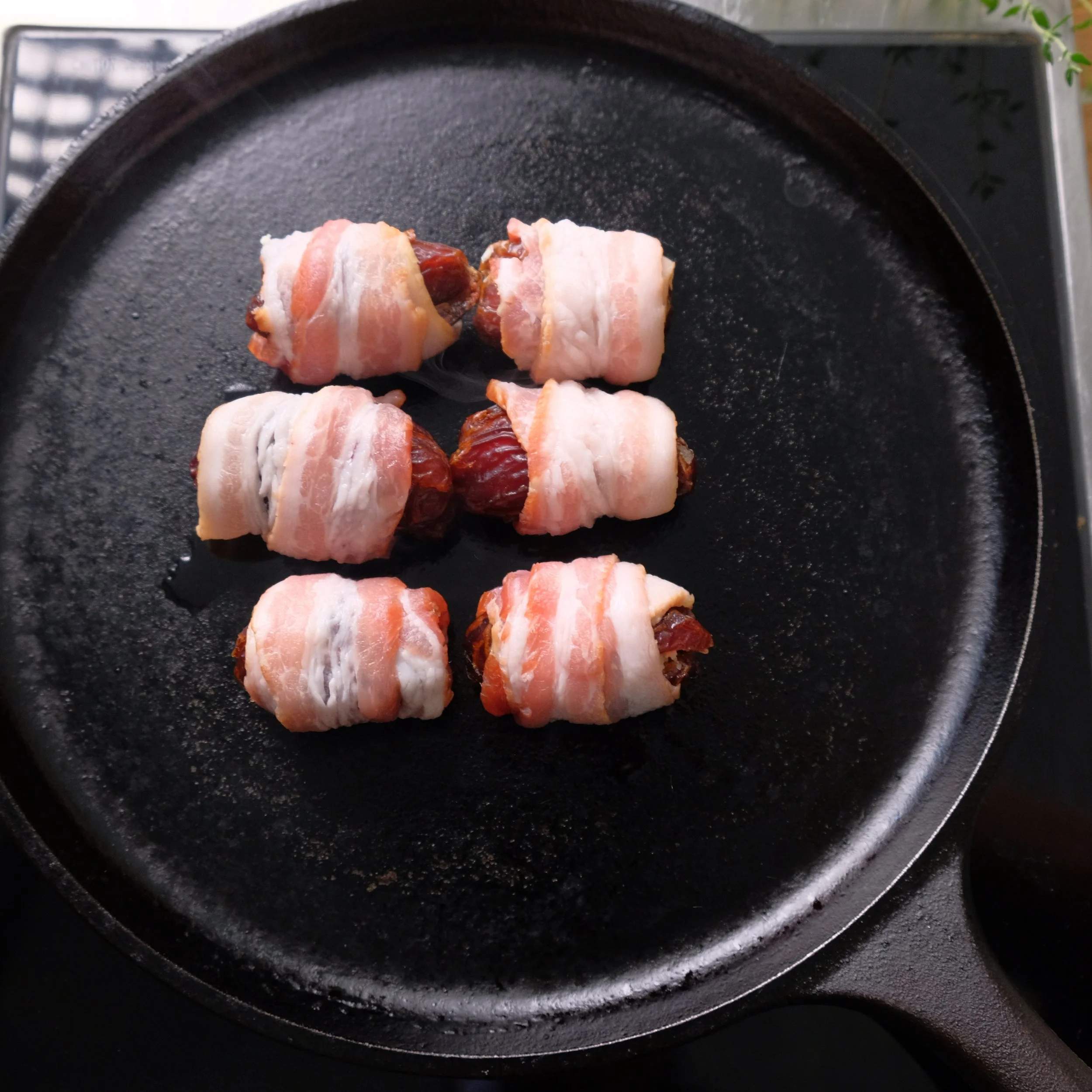 Six bacon-wrapped dates on a black skillet, ready for cooking.