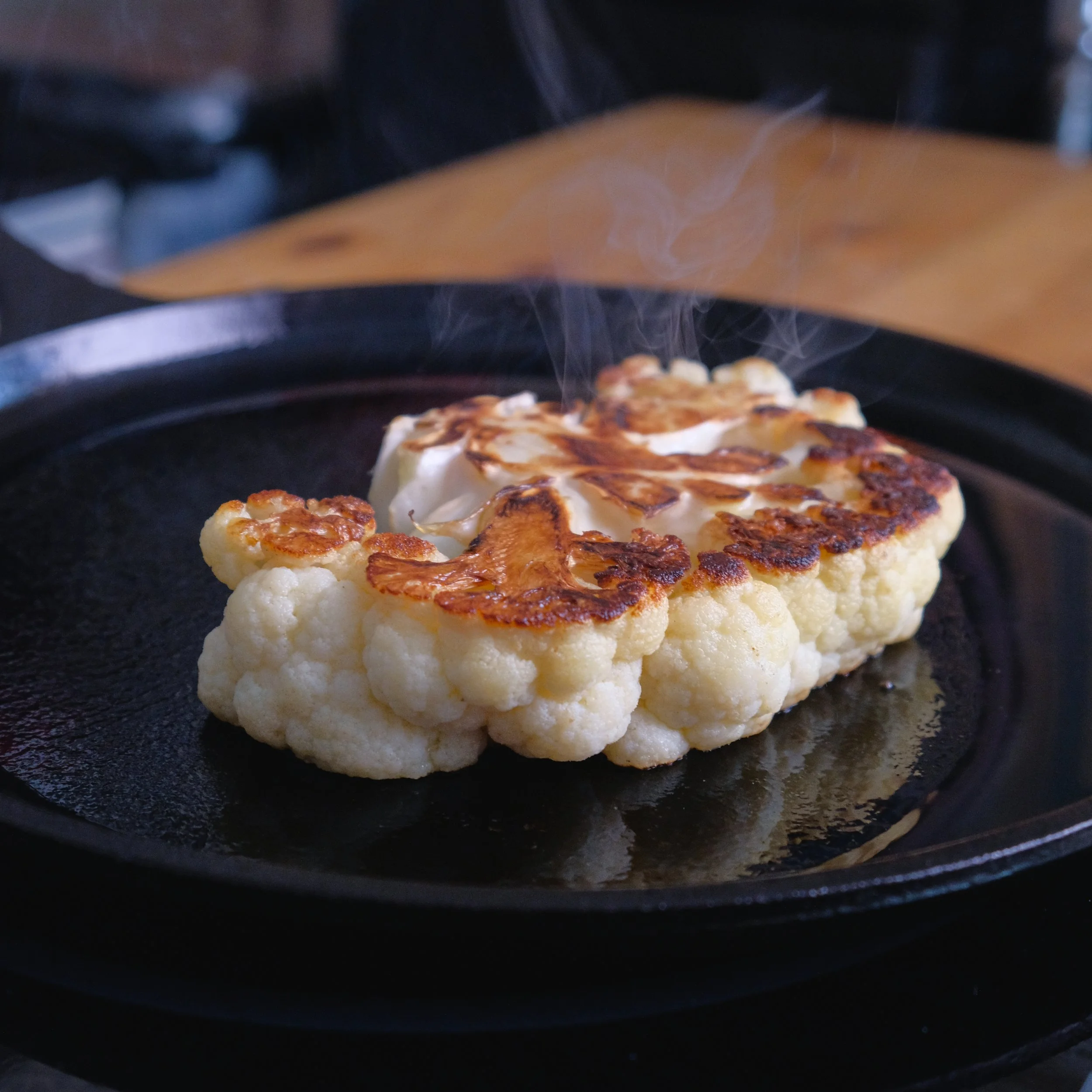 A head of cauliflower being cooked on a black cast iron skillet, with some parts browned and steaming.