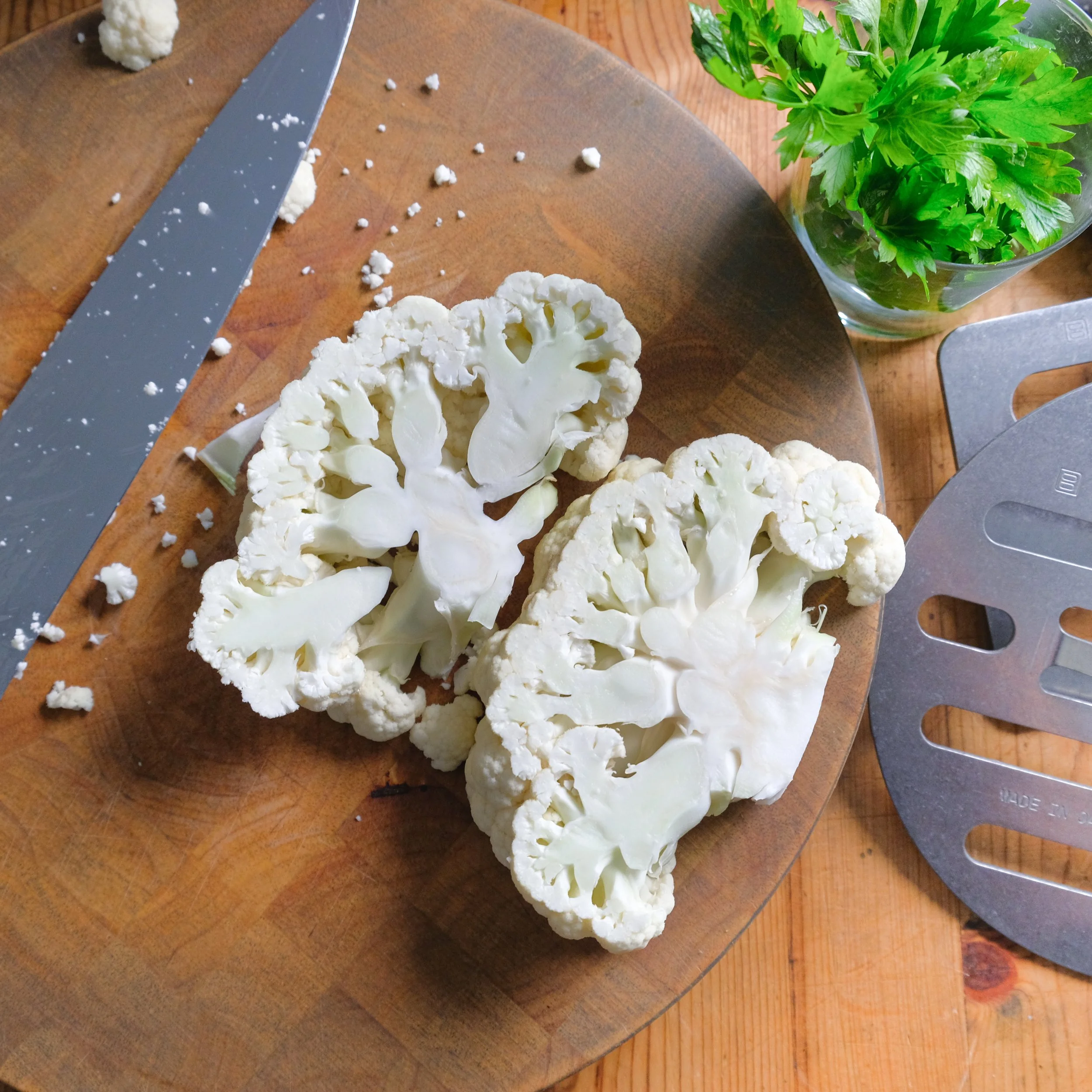 Fresh cauliflower cut into florets on a wooden cutting board, with a knife nearby, a glass bowl of fresh parsley, and metal vegetable slicer.