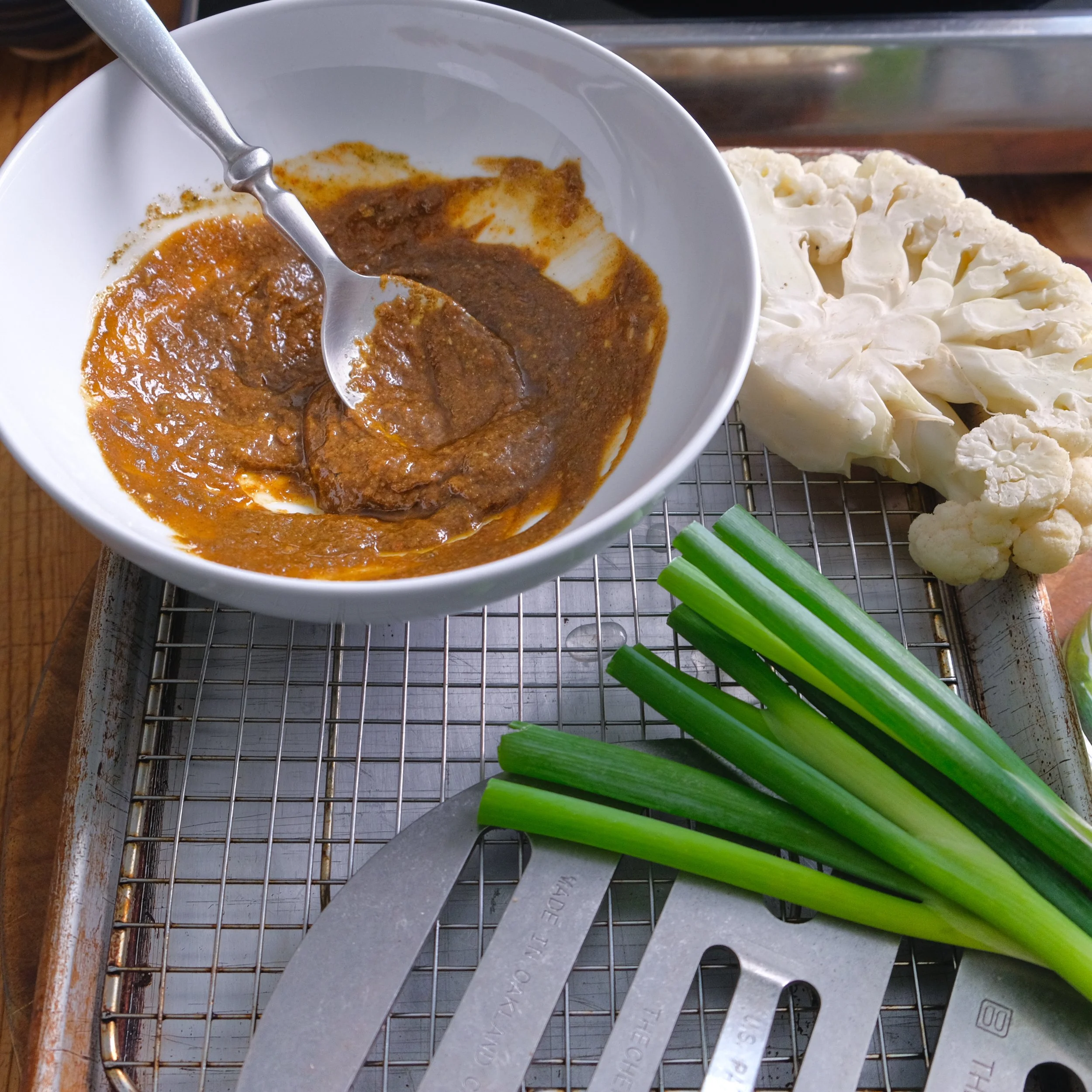A white bowl with a spoon containing thick reddish-brown curry sauce, placed on a metal grid tray. Nearby are fresh green onions, cauliflower florets, and a large piece of cauliflower head. There are also metal tongs resting on the tray.