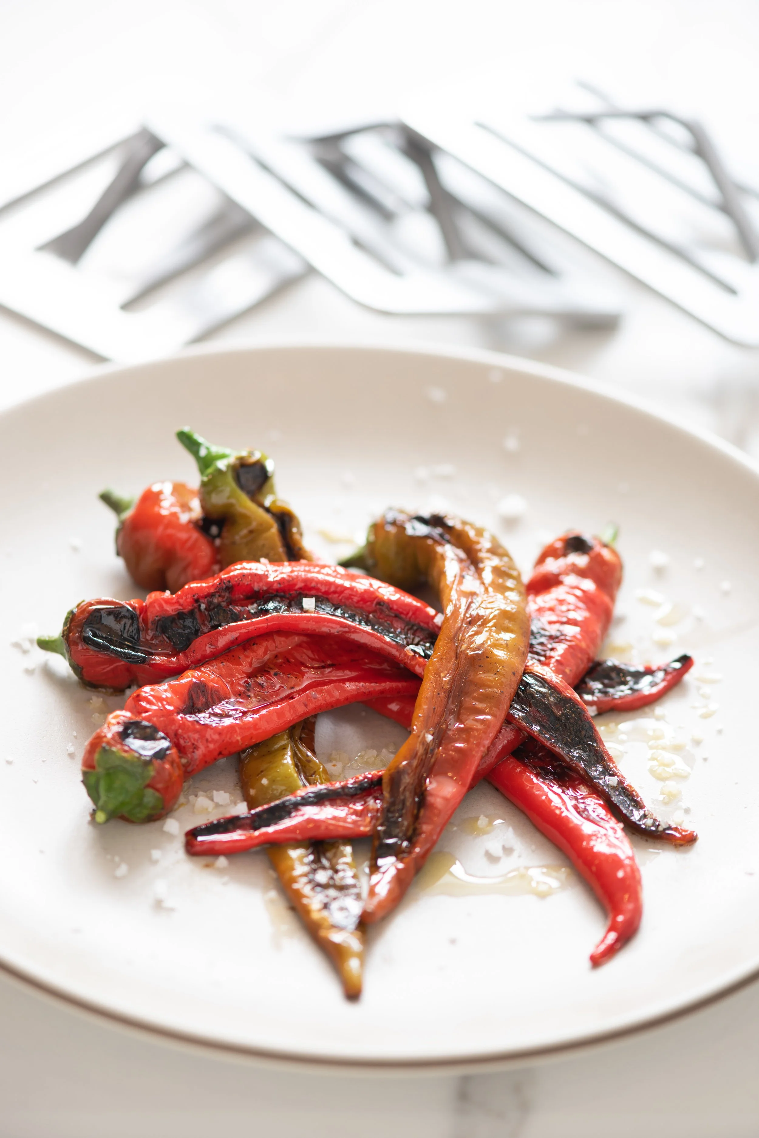Charred colorful peppers on a white plate with salt and blurred utensils in background.