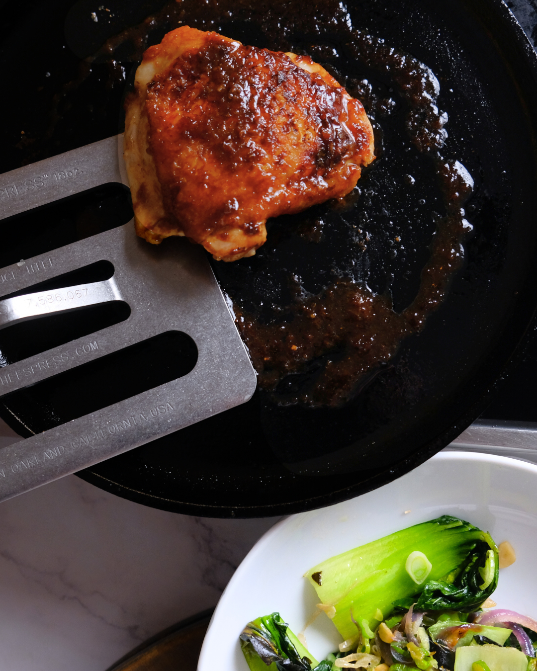 A piece of chicken with barbecue sauce in a frying pan and a bowl of salad with greens and onions next to it.