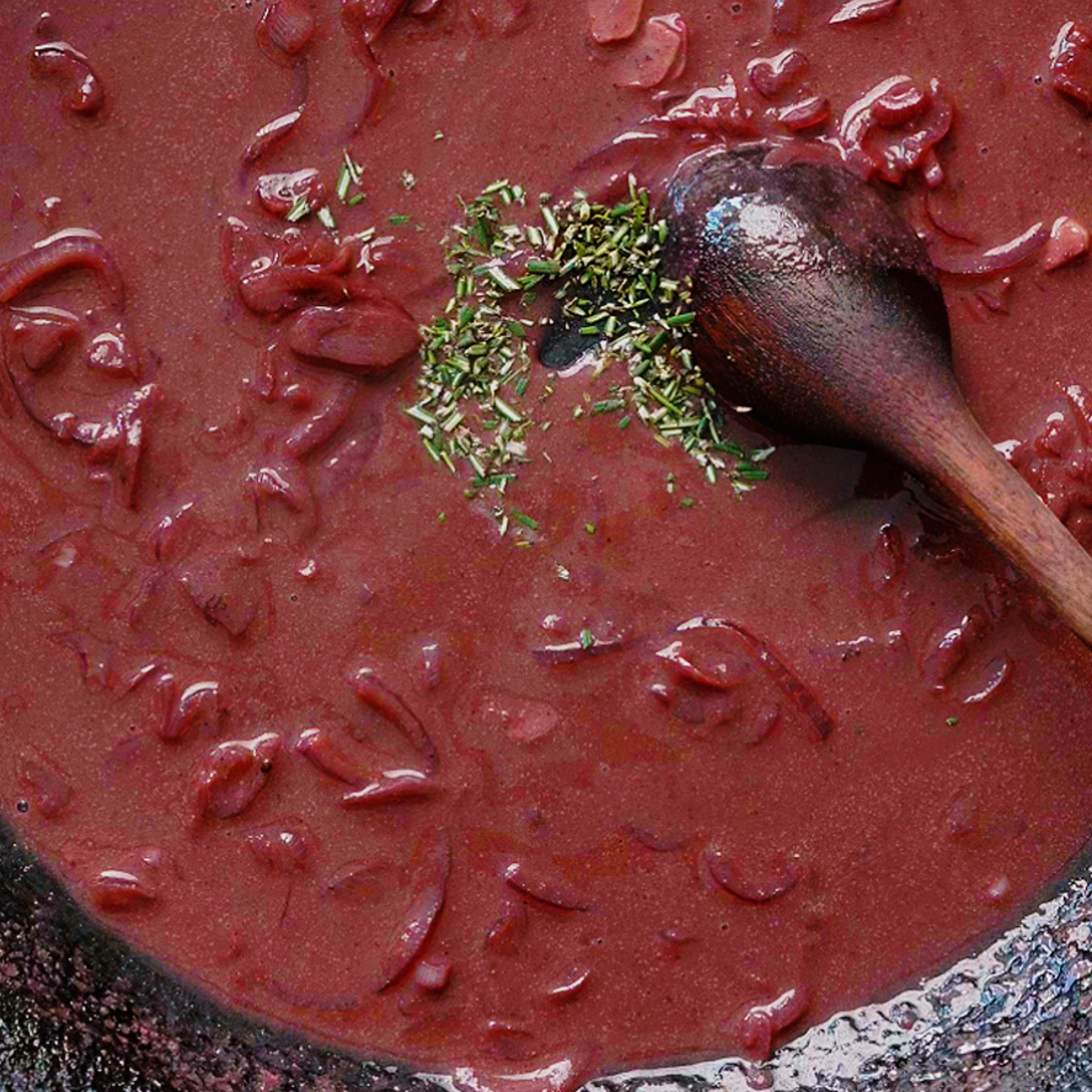 A pot of red pasta with sliced peppers, topped with chopped herbs and a wooden spoon.