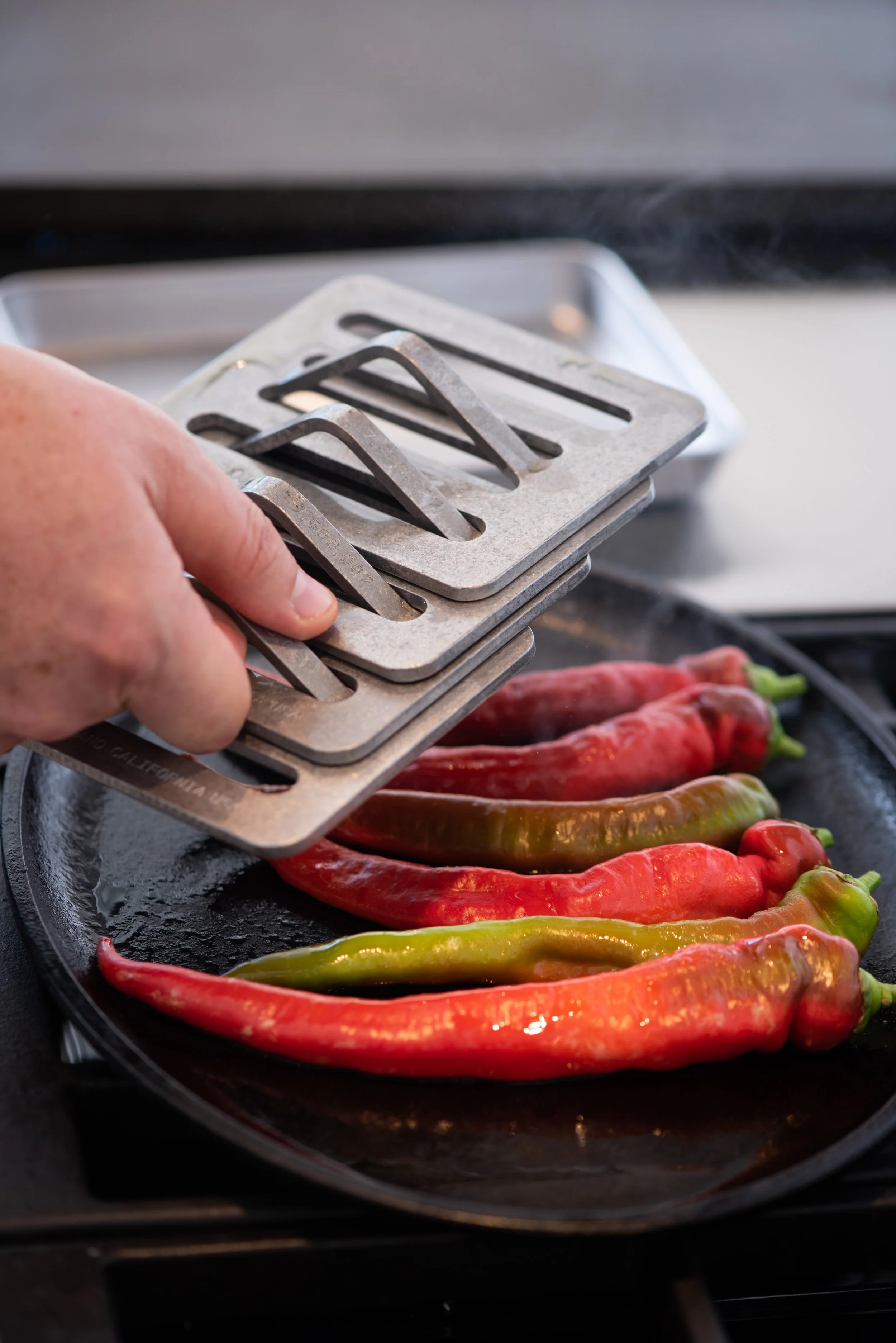 Hand placing metal chili pepper griddle press on a tray of red and green chili peppers on a stovetop.