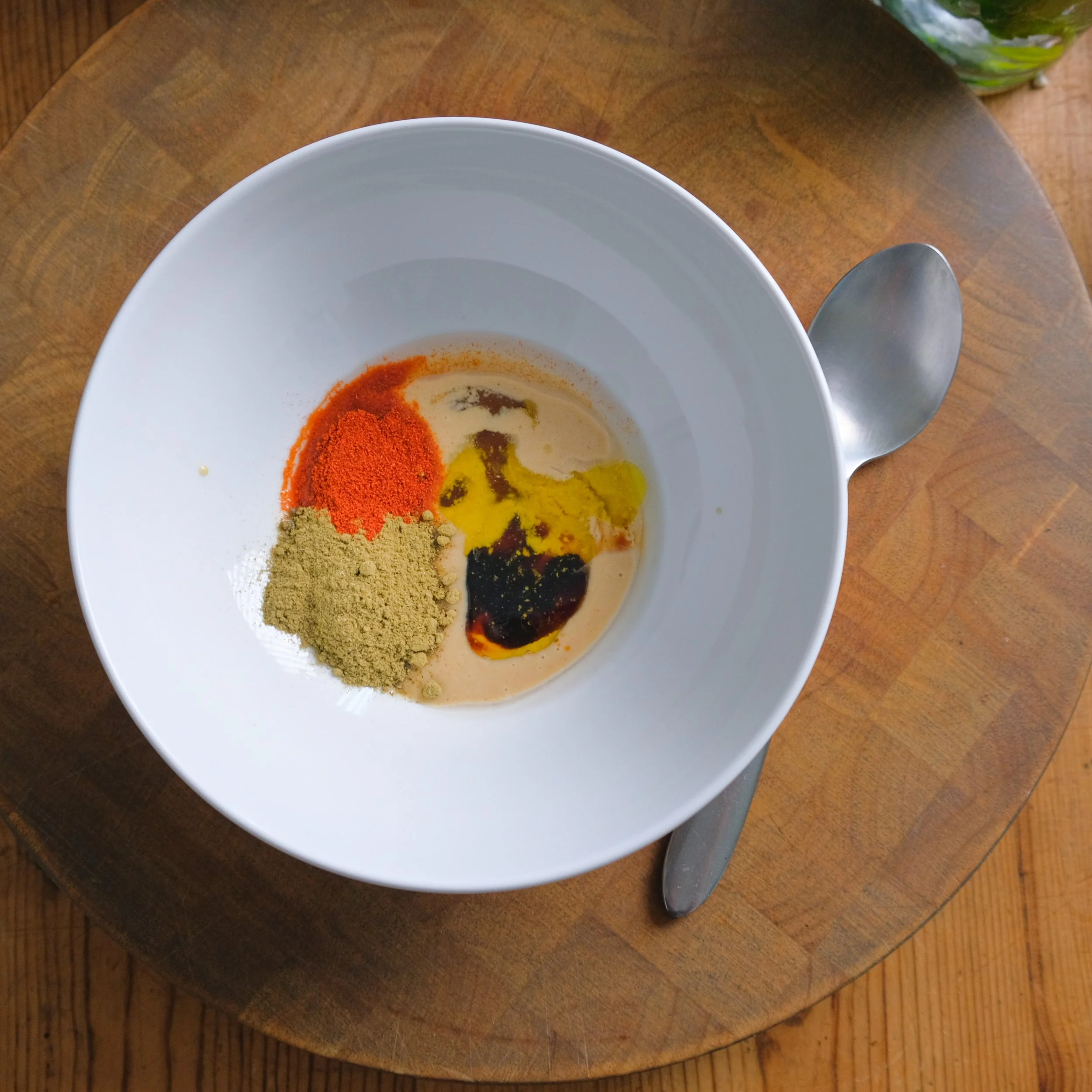 A white bowl containing various spices and liquids, including red, green, and dark brown powders and sauces, on a wooden table with a spoon beside it.