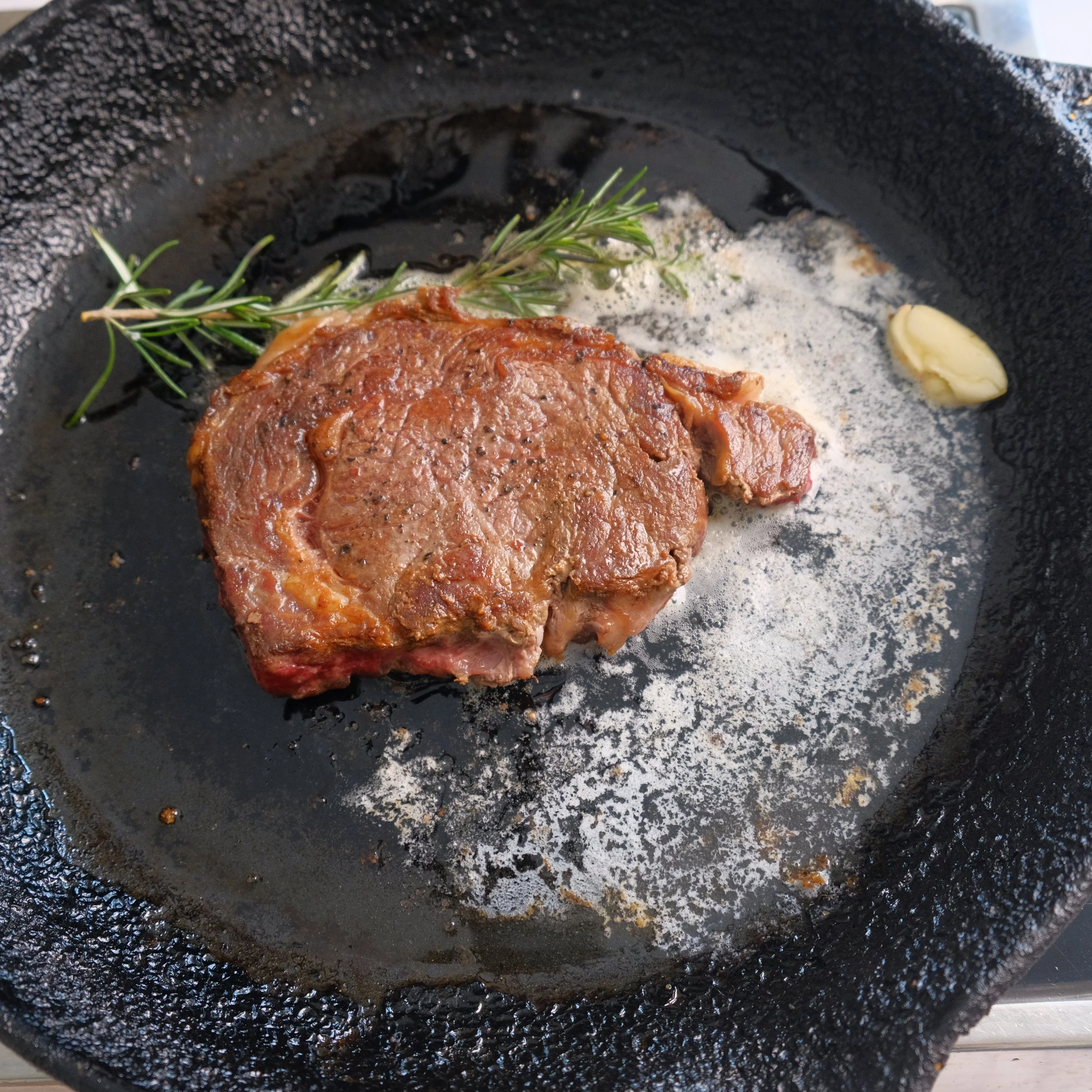 A raw steak seasoned with herbs, butter, and garlic in a cast iron skillet with a sprig of rosemary and a clove of garlic.