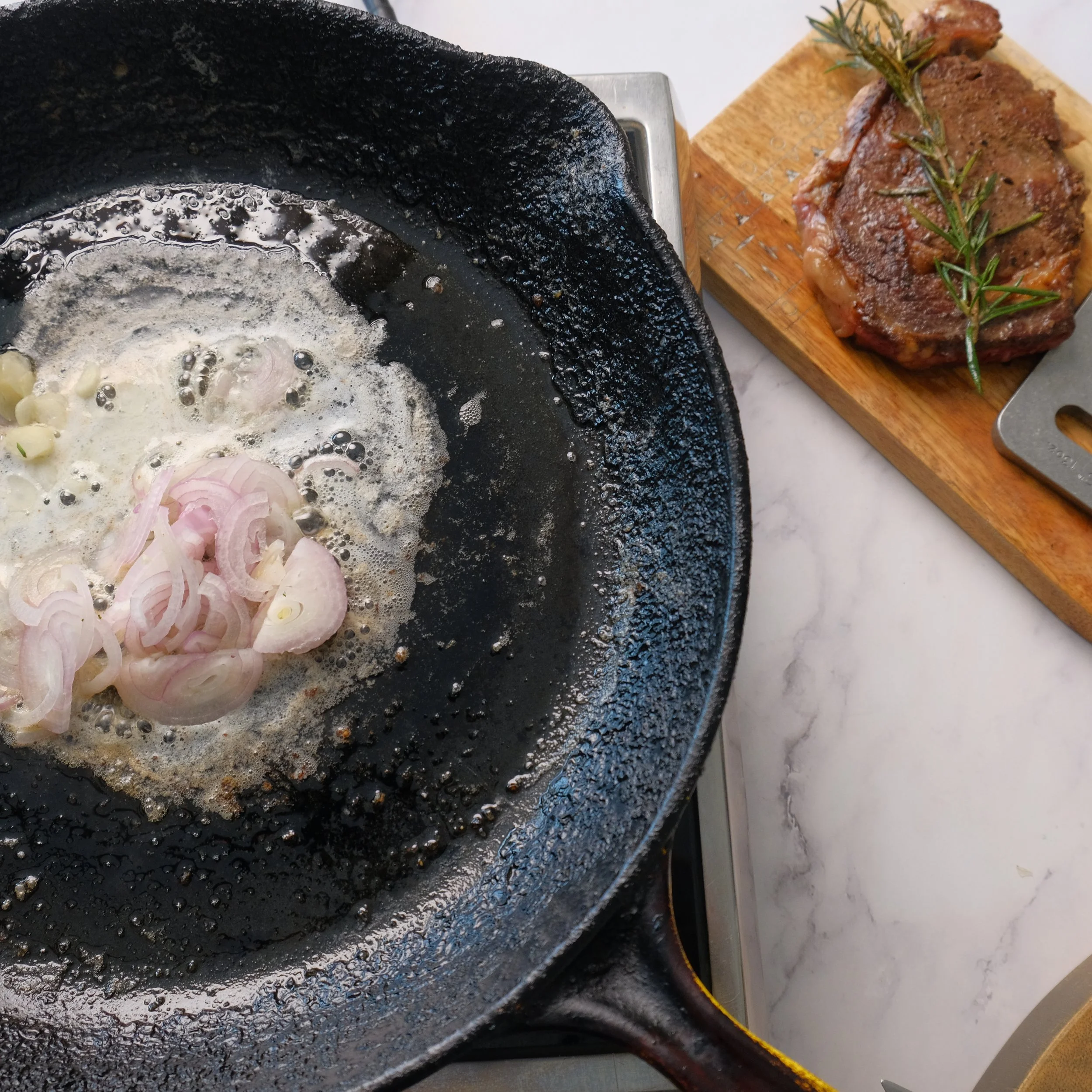 Cooking onions in a cast iron skillet on a stove, with raw steak seasoned with herbs on a wooden cutting board nearby.