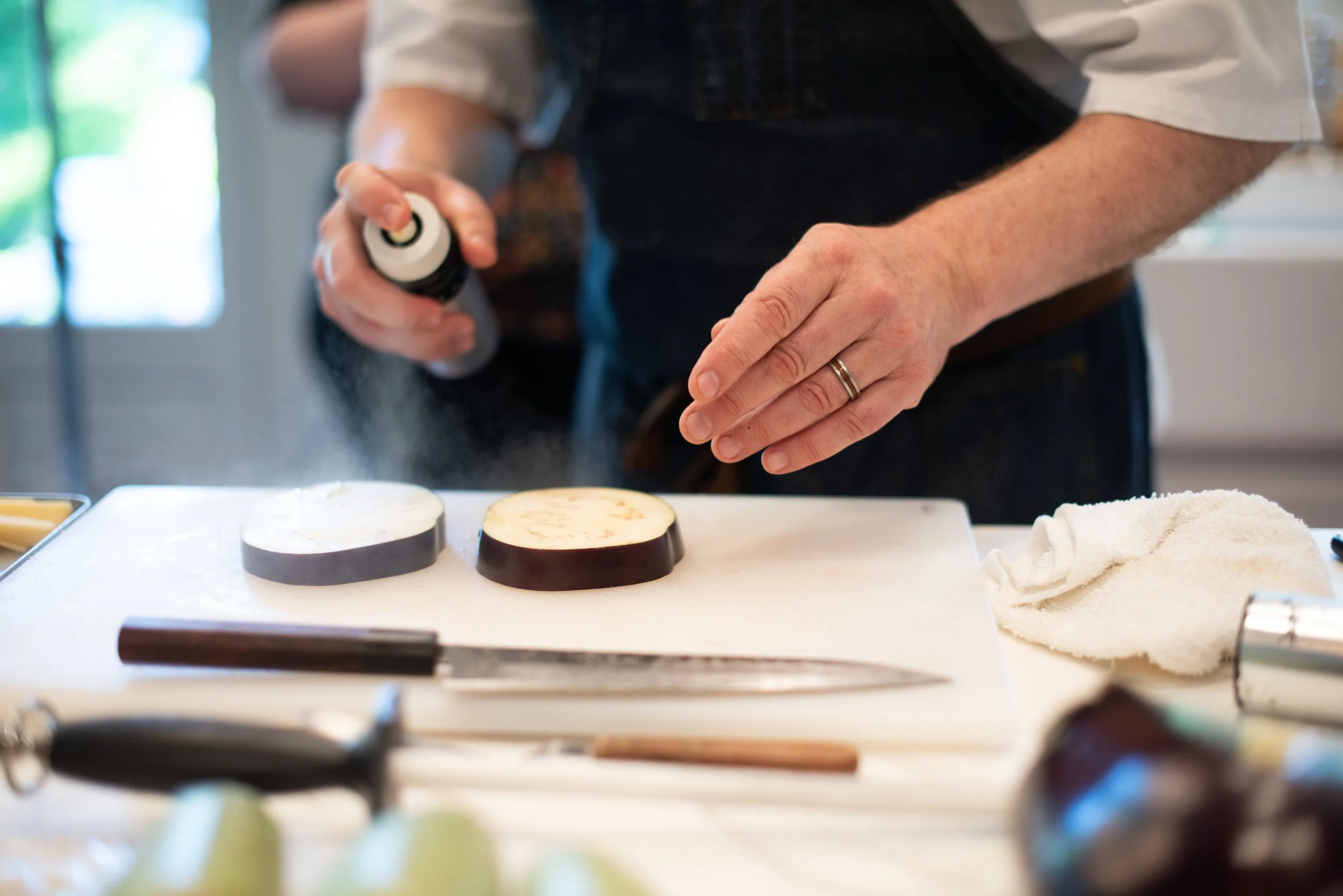 A person sprinkles seasoning onto two slices of eggplant on a white cutting board in a kitchen.
