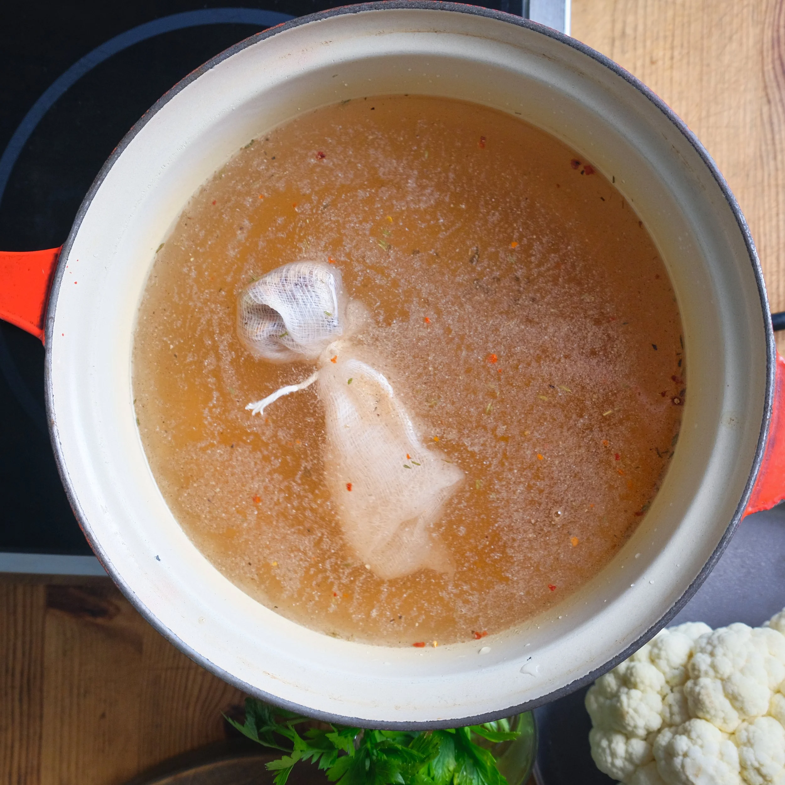 Pot of broth with a tea bag inside, resting on a stovetop with cauliflower and fresh herbs nearby.