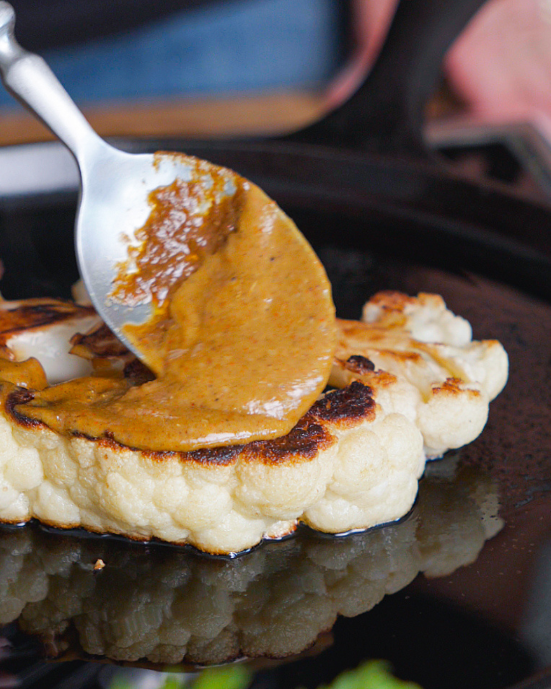 A spoon pouring curry sauce over roasted cauliflower on a black griddle.