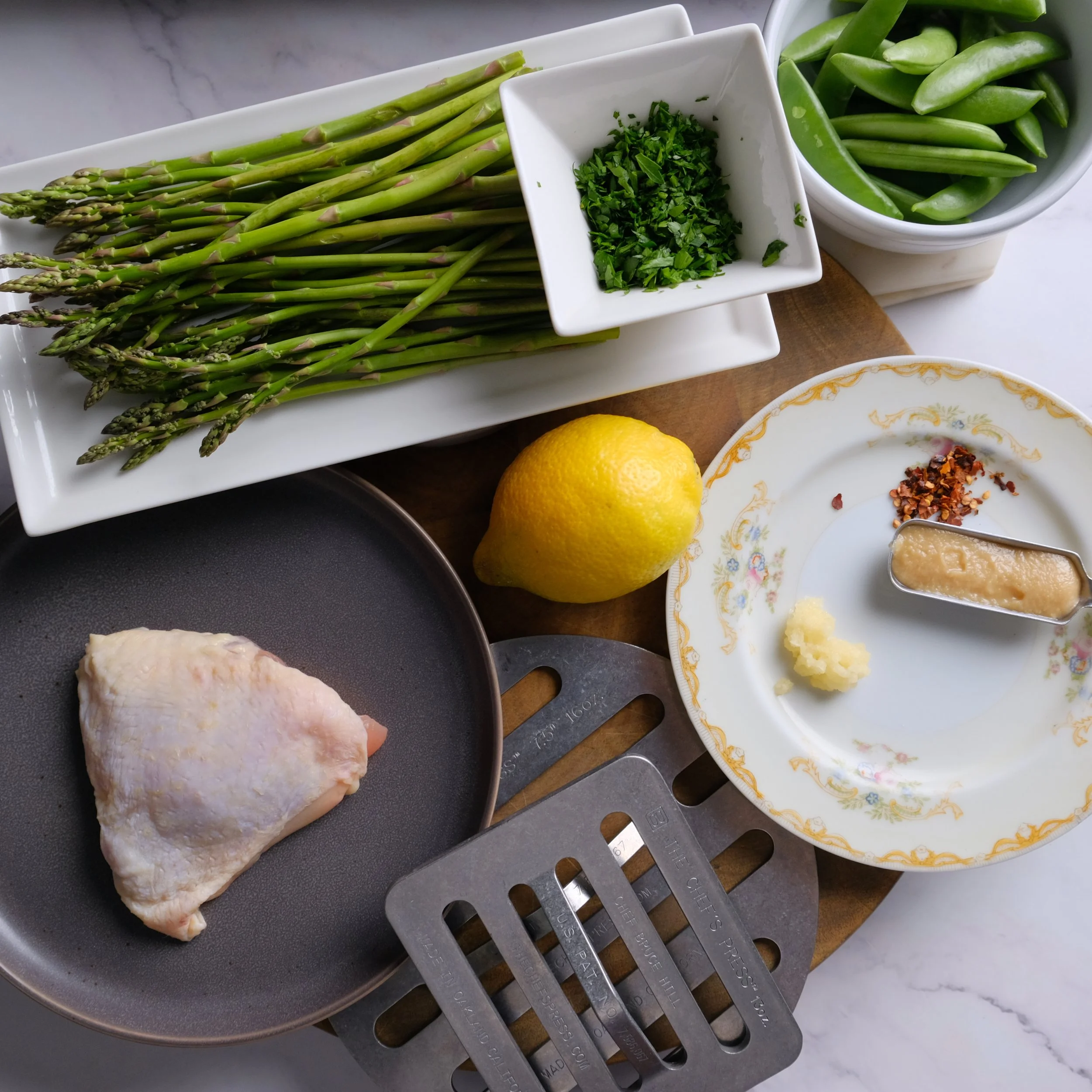 Ingredients for a meal including a bunch of asparagus, chopped herbs, snow peas, a lemon, a chicken thigh in a frying pan, minced garlic, red pepper flakes, and ginger on a decorative plate, with a metal grater and a lemon zester.