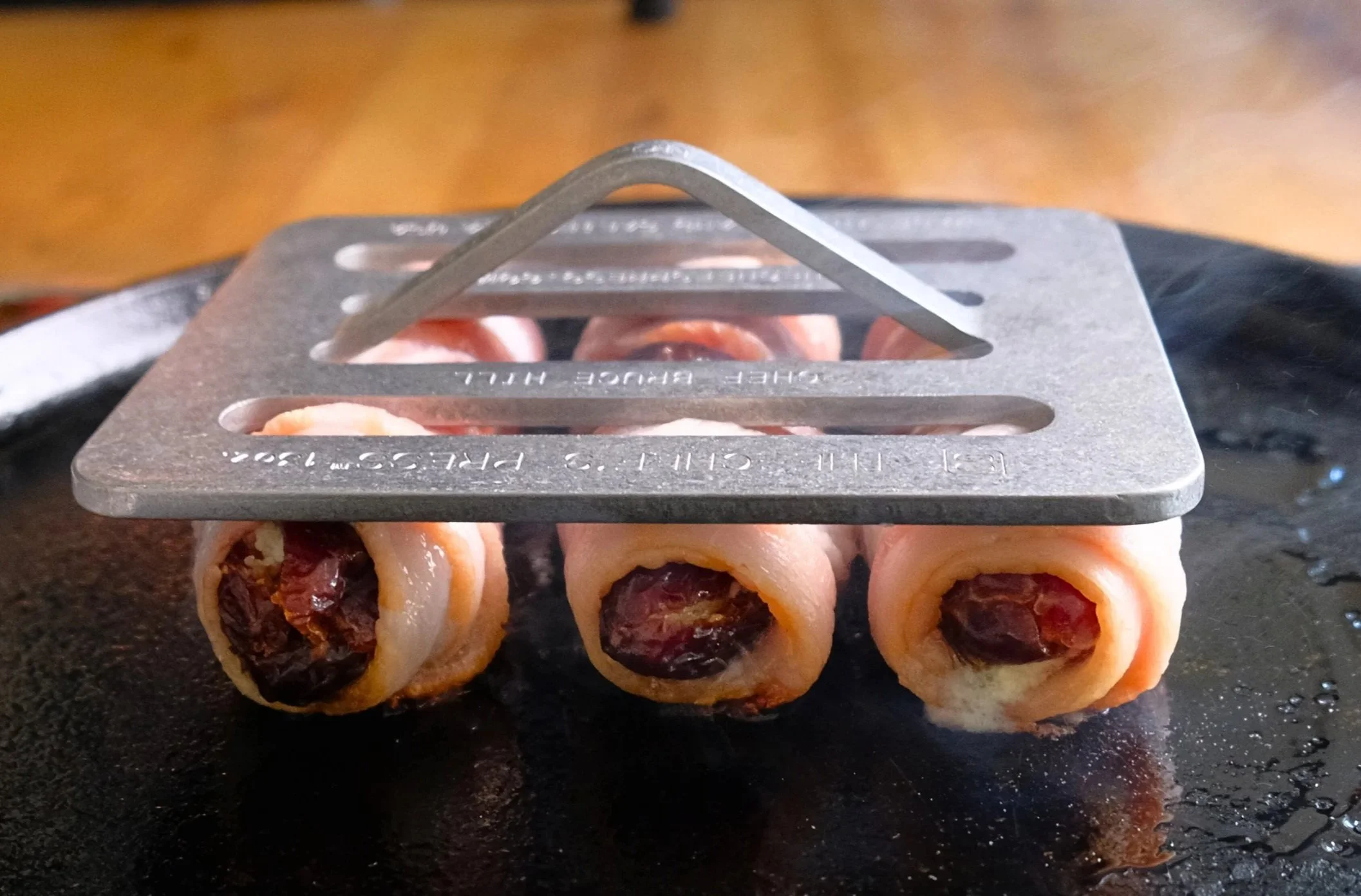 Rolled raw bacon-wrapped stuffed dates on a black surface with a metal bacon press on top.
