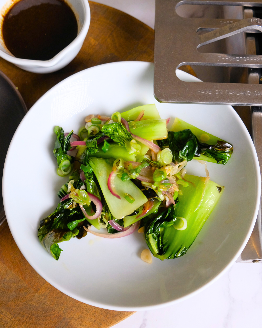 Fresh green salad with sliced cucumbers, purple onions, and leafy greens on a white plate, with a small bowl of dark dressing and a stovetop burner nearby.