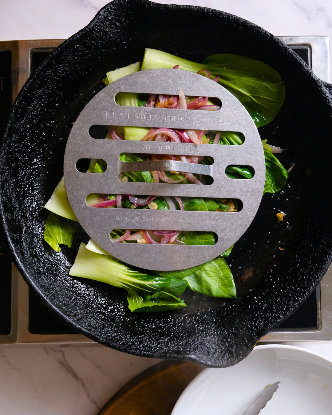 Fresh vegetables in a black cast iron skillet topped with a metal grill, including bok choy, spinach, and red onions, on a stove.