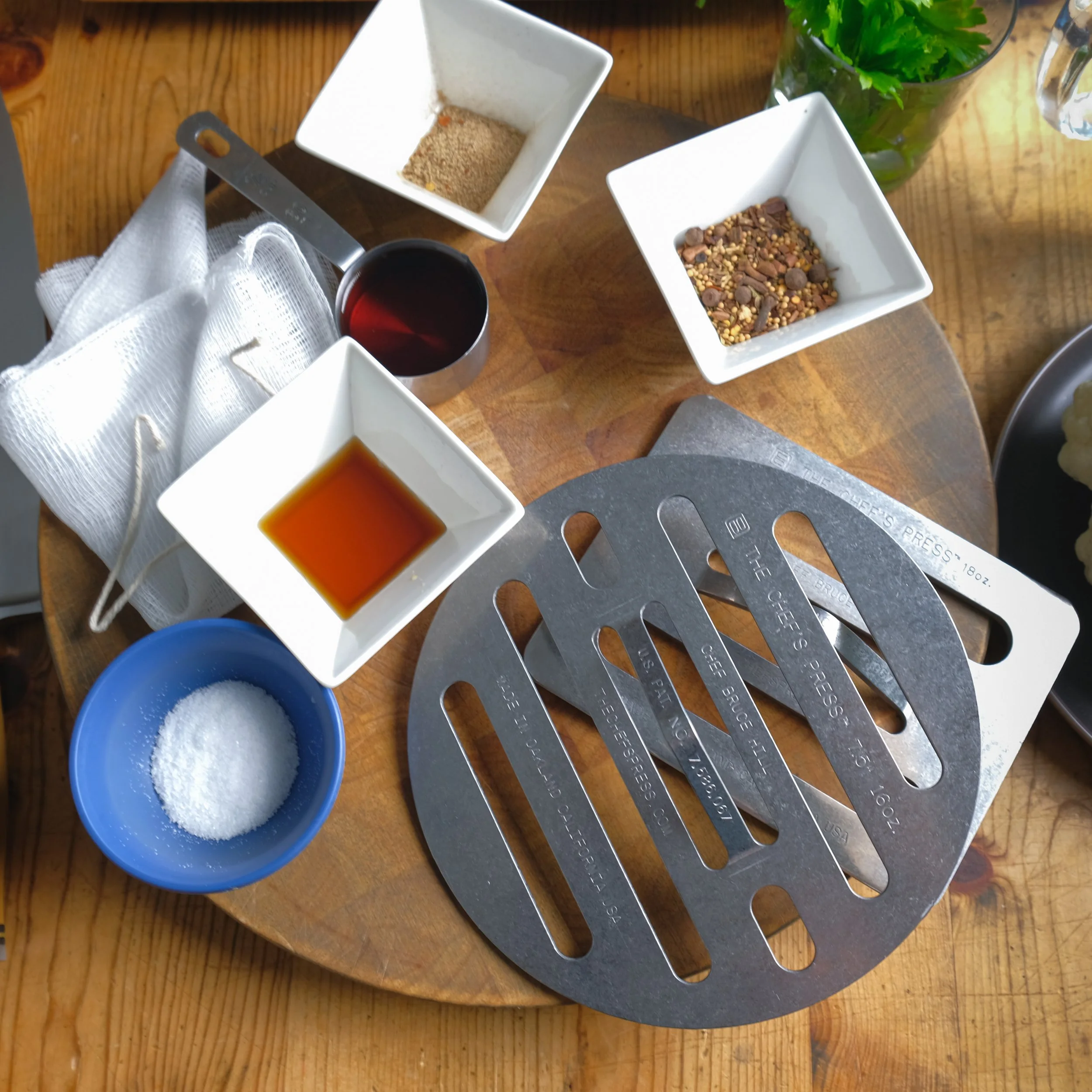 A round wooden table with four white square bowls containing seasonings and sauces, a small blue bowl of salt, a small metal cup of soy sauce, and a small metal cup of a reddish liquid, with a metal cheese slicer and some green leafy herbs.