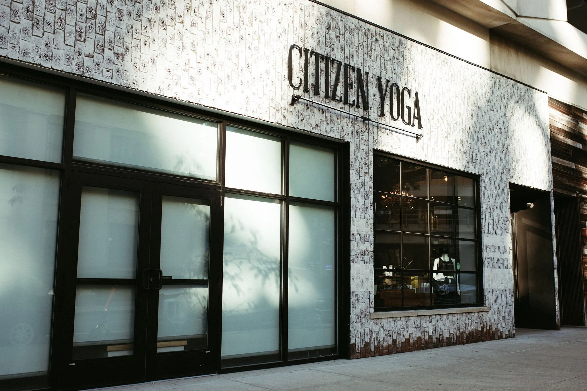 Exterior view of Citizen Yoga studio in a building with a brick facade and large glass windows, one of which displays a rainbow pride flag.