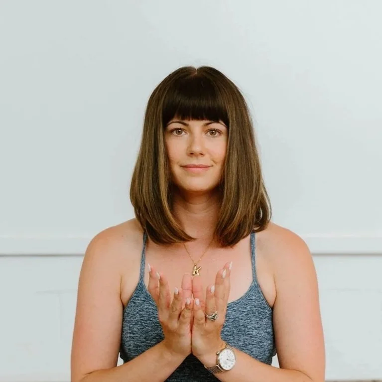 Close-up of a young woman with long brown hair and light brown eyes, outdoors in natural light, with a blurred gray wall in the background.