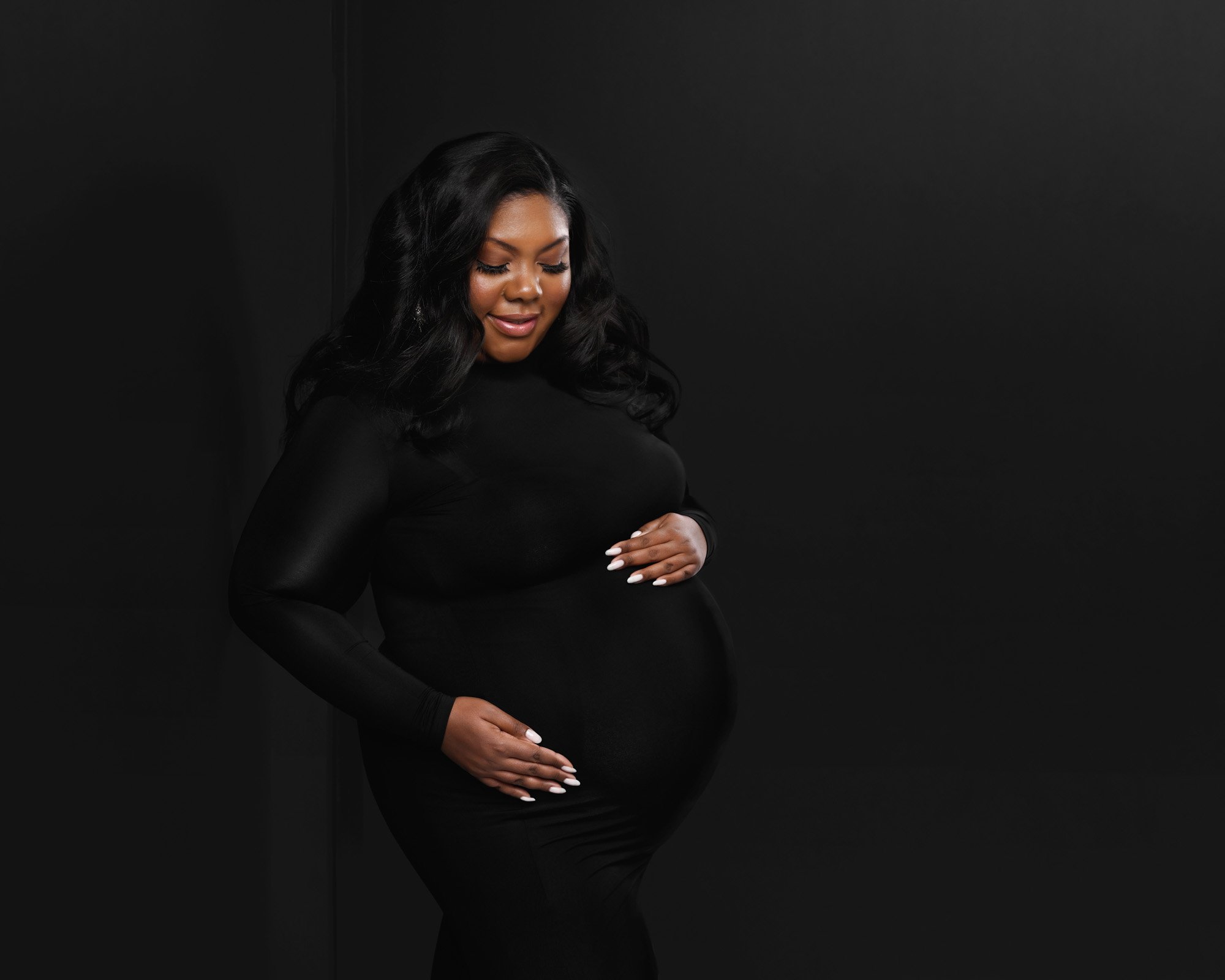 Pregnant woman in black dress holding her belly against a black background.