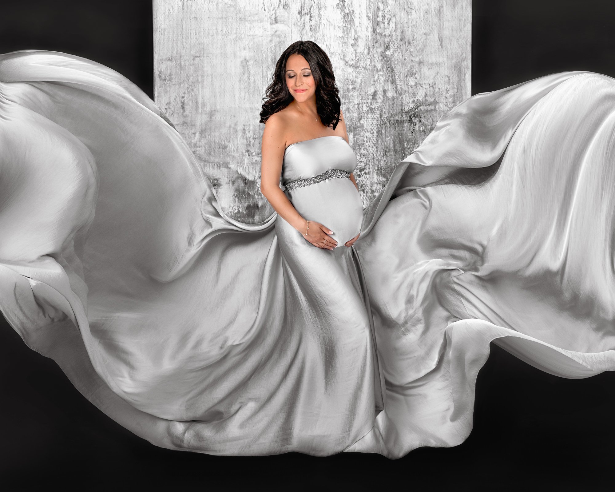 Pregnant woman in flowing white gown holding belly with gray background.