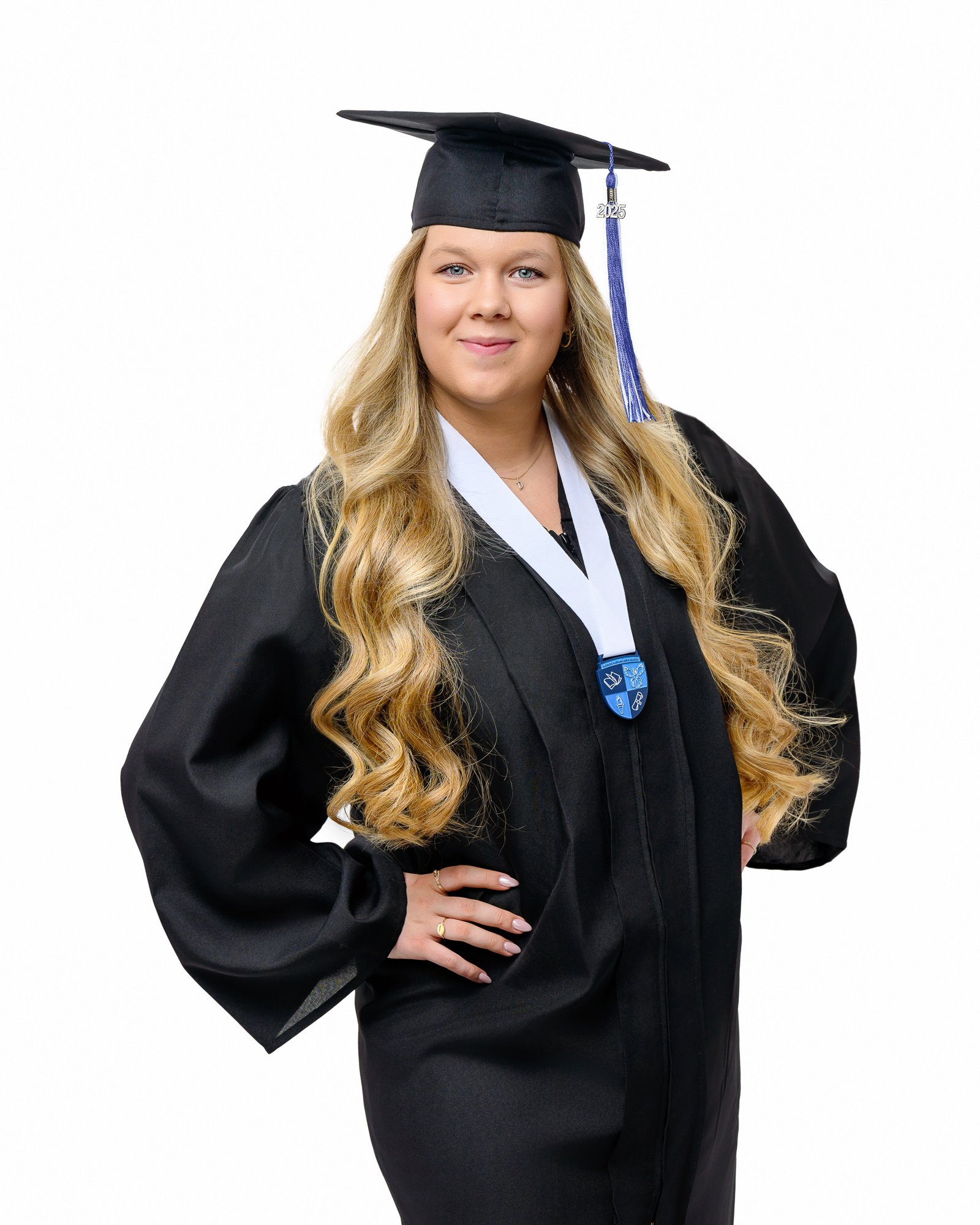 Child in blue graduation cap and gown with yellow sash, leaning against a wall, smiling, outdoors with greenery in the background.