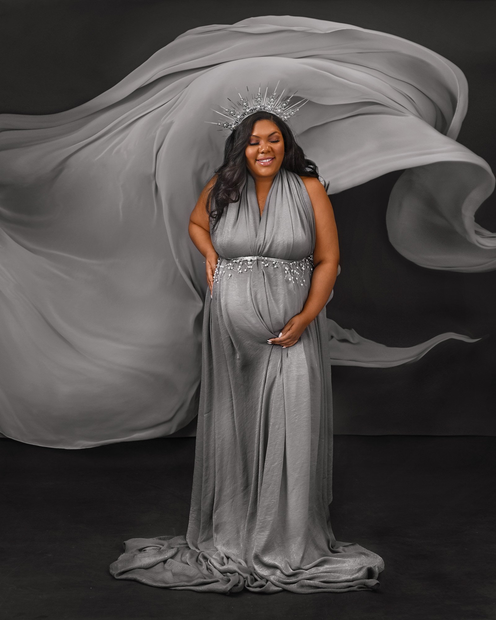Pregnant woman in a gray dress with a crown, standing against a flowing fabric background, posing with a hand on her belly.