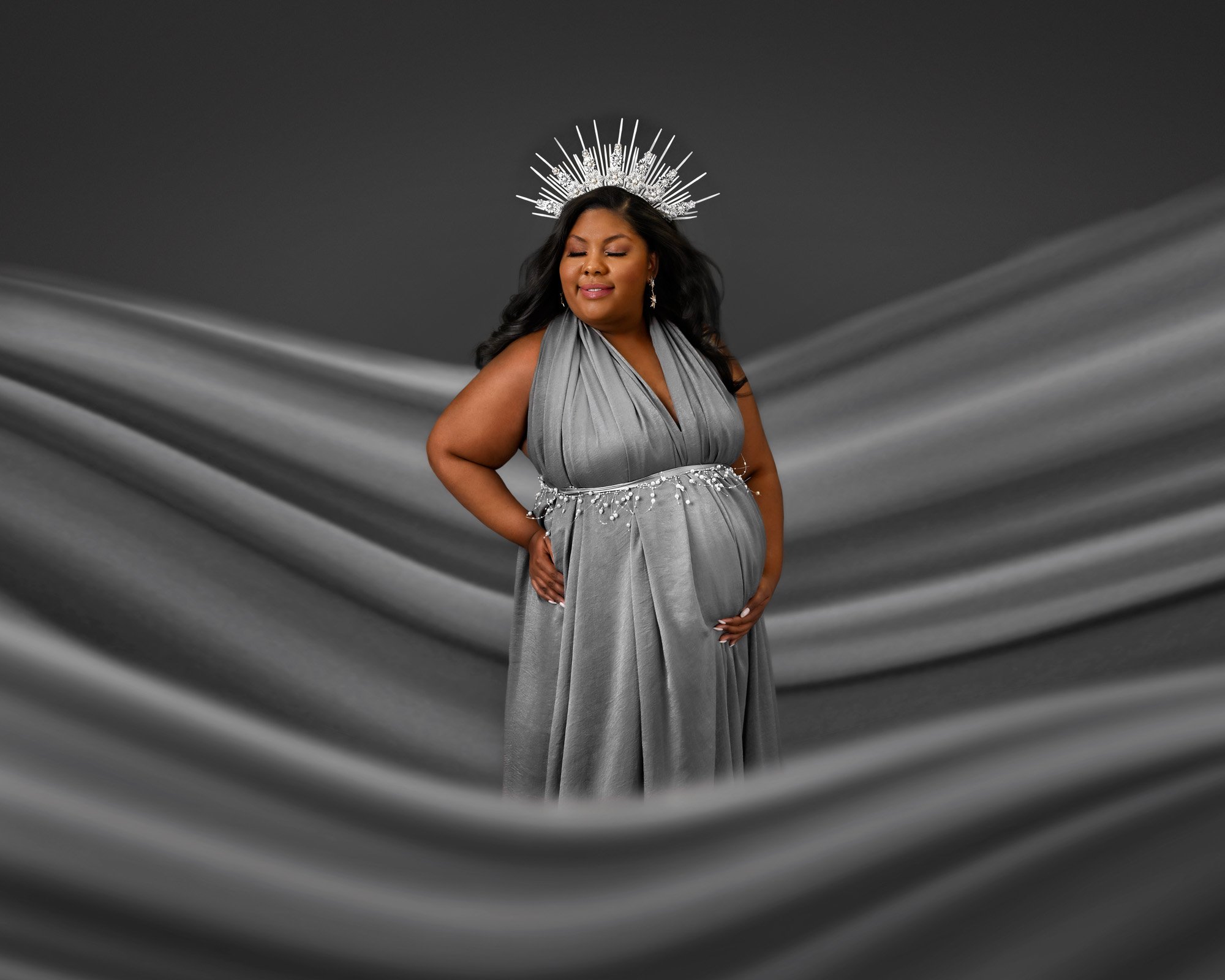 Pregnant woman in gray dress with a crown against a wavy fabric background.
