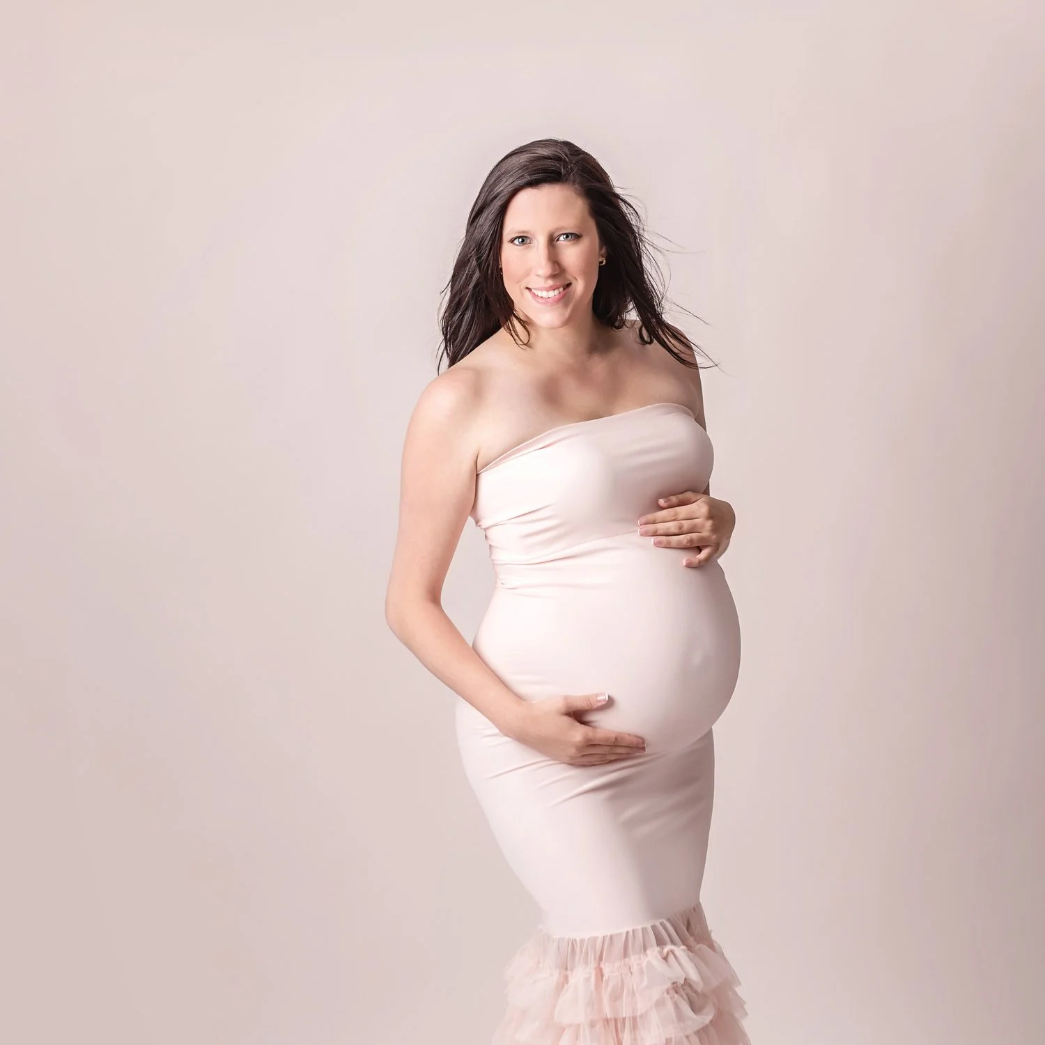 Pregnant woman in a strapless pink dress holding her belly, smiling against a neutral background.