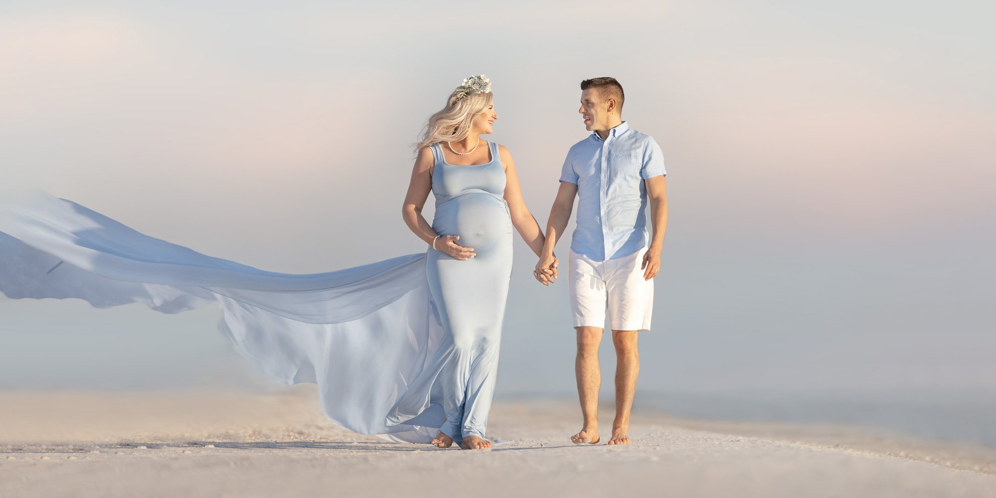 Pregnant woman in blue dress and man holding hands walking on beach