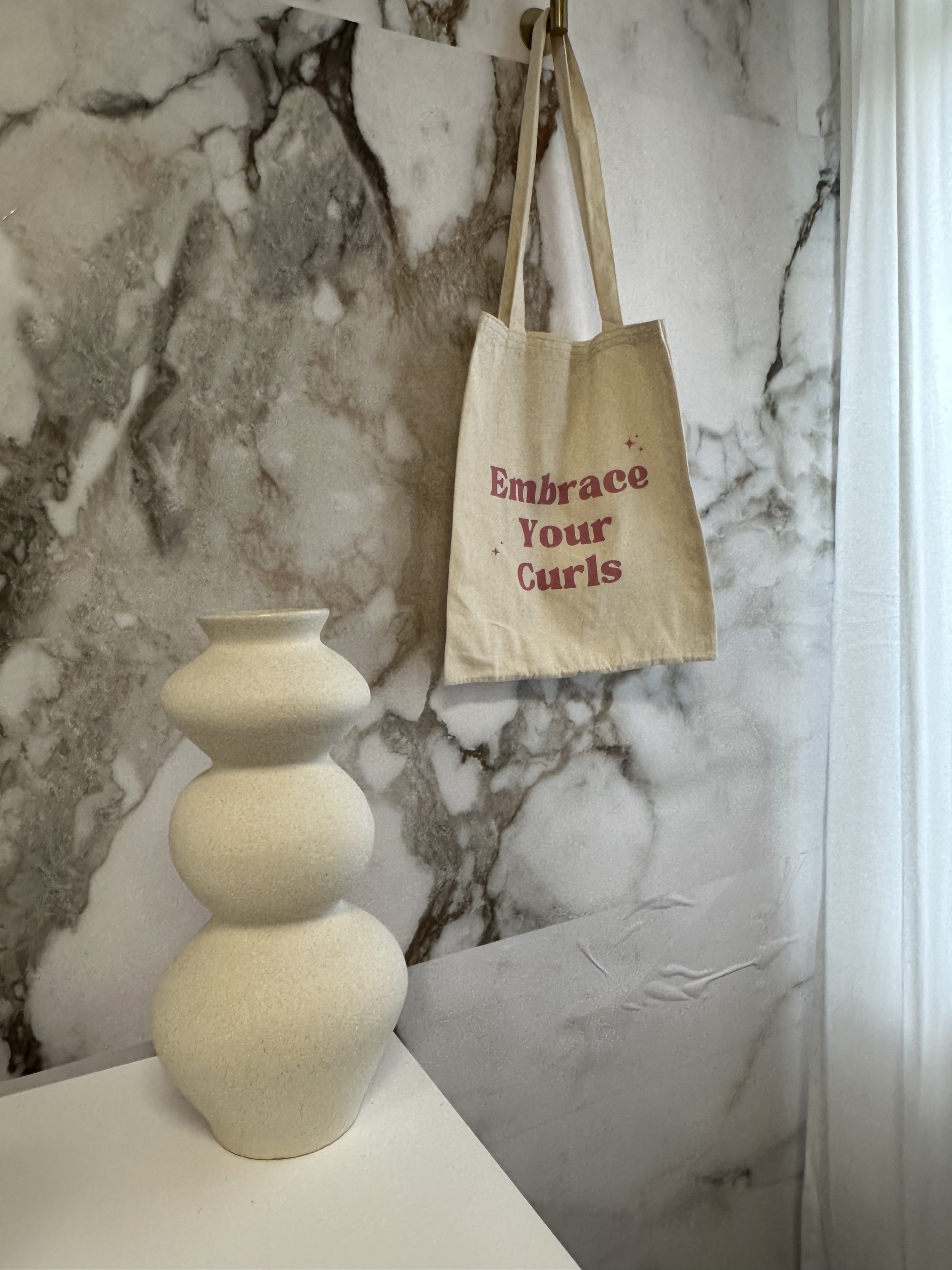 A beige ceramic vase with three bulbous sections placed on a white surface, with a marble wall in the background and a beige tote bag hanging on a hook that says 'Embrace Your Curls' in burgundy text.