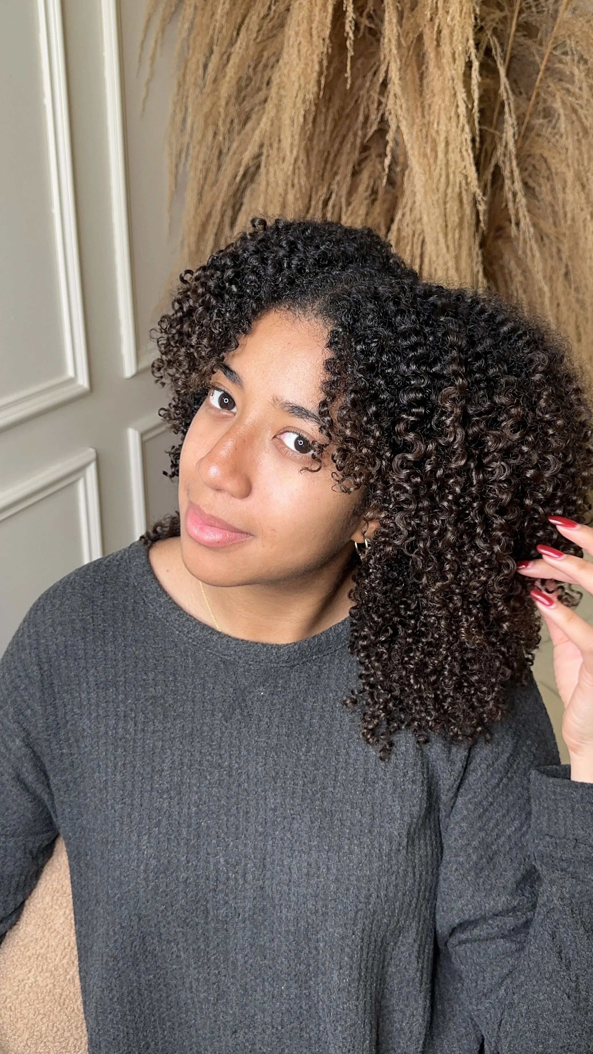A woman with natural curly hair touching her hair, wearing a black top, earrings, and a subtle smile, with a background of dried plants and white paneled walls.