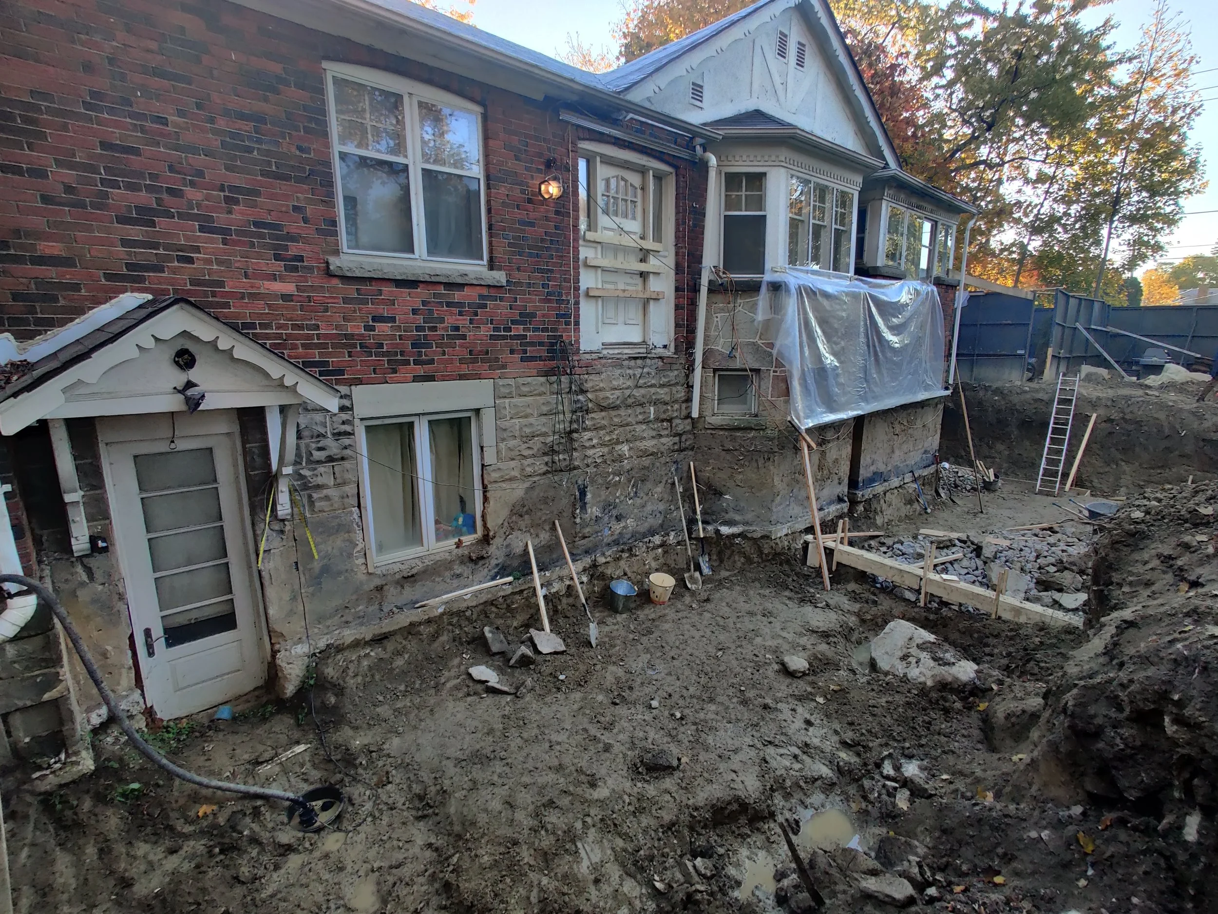 The back of a house under renovation with exposed foundation. The house has brick walls and a white bay window. There is a small wooden structure with a gabled roof and a decorative trim. A plastic sheet covers part of the porch, and there is a ladde