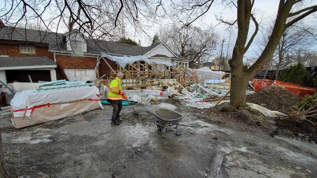 Construction worker in a high-visibility jacket working outdoors on a construction site in front of a partially demolished building with scaffolding and construction materials.