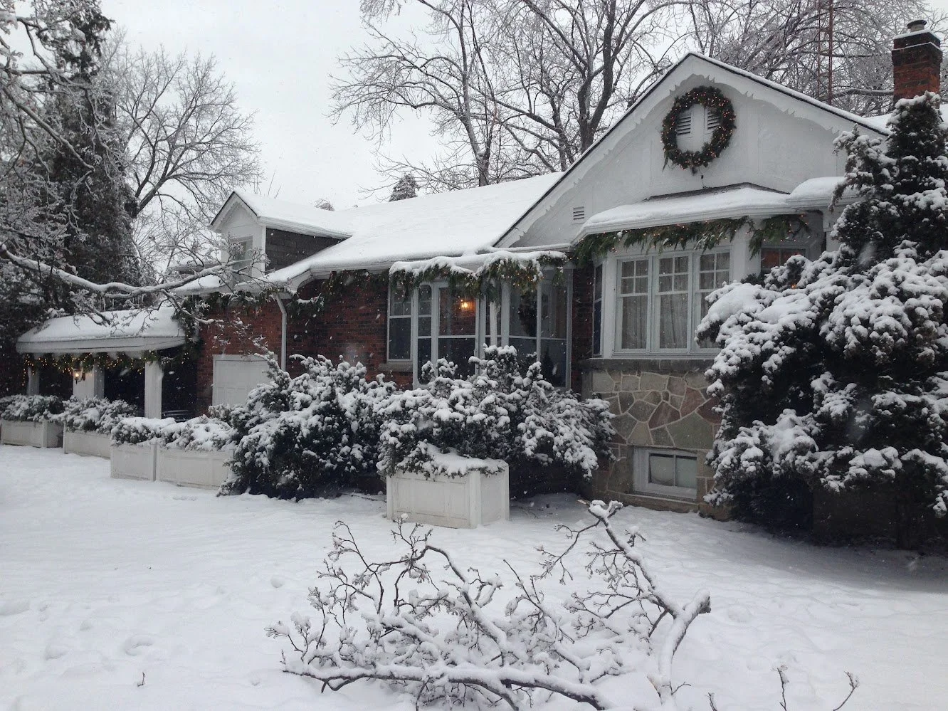 A house covered in snow, decorated with a Christmas wreath and garland, surrounded by snow-covered trees and bushes.