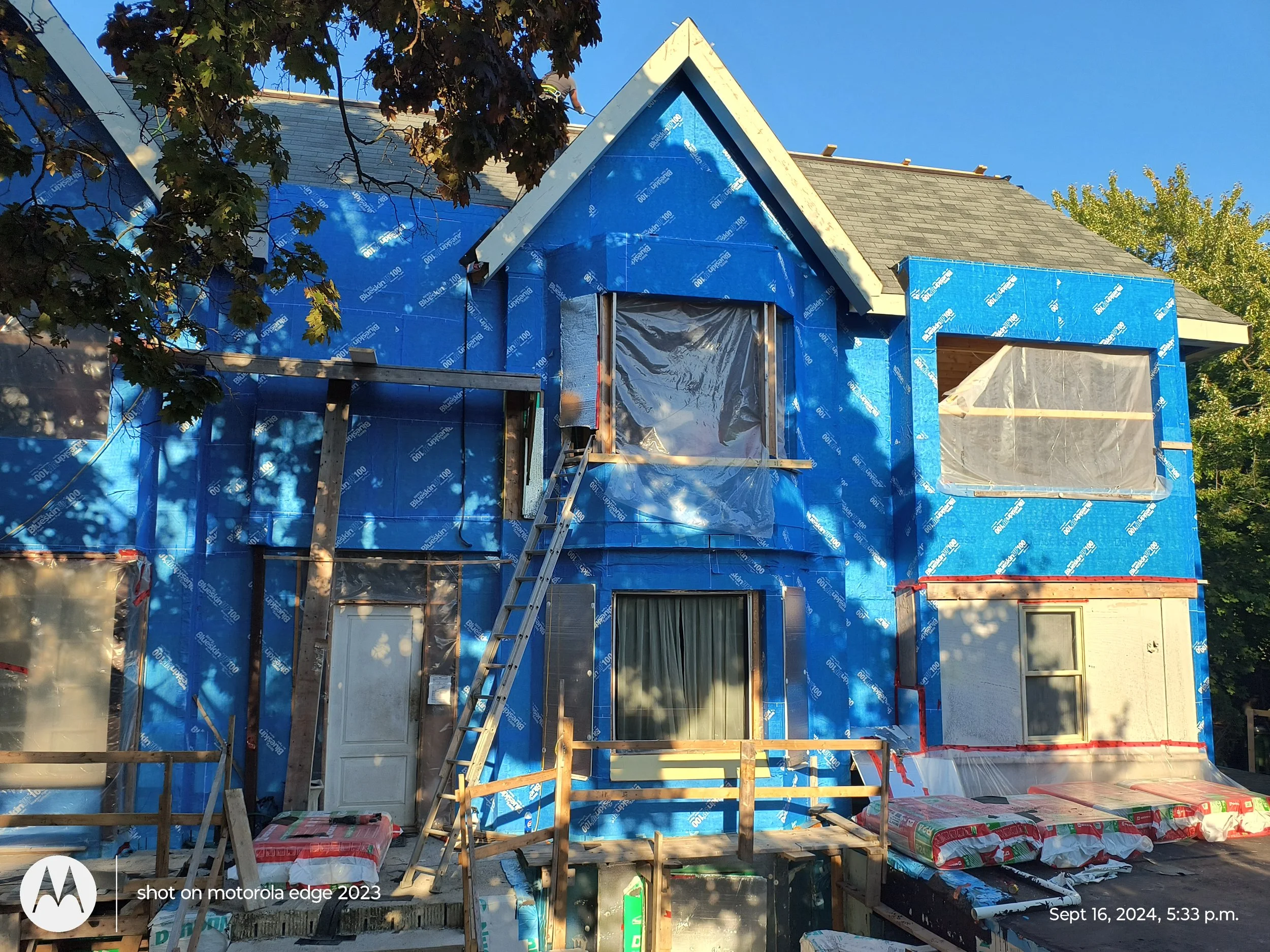 A house under construction with a blue weather-resistant barrier on the exterior walls, with wooden framing and tools visible, and scaffolding around the building at sunset.